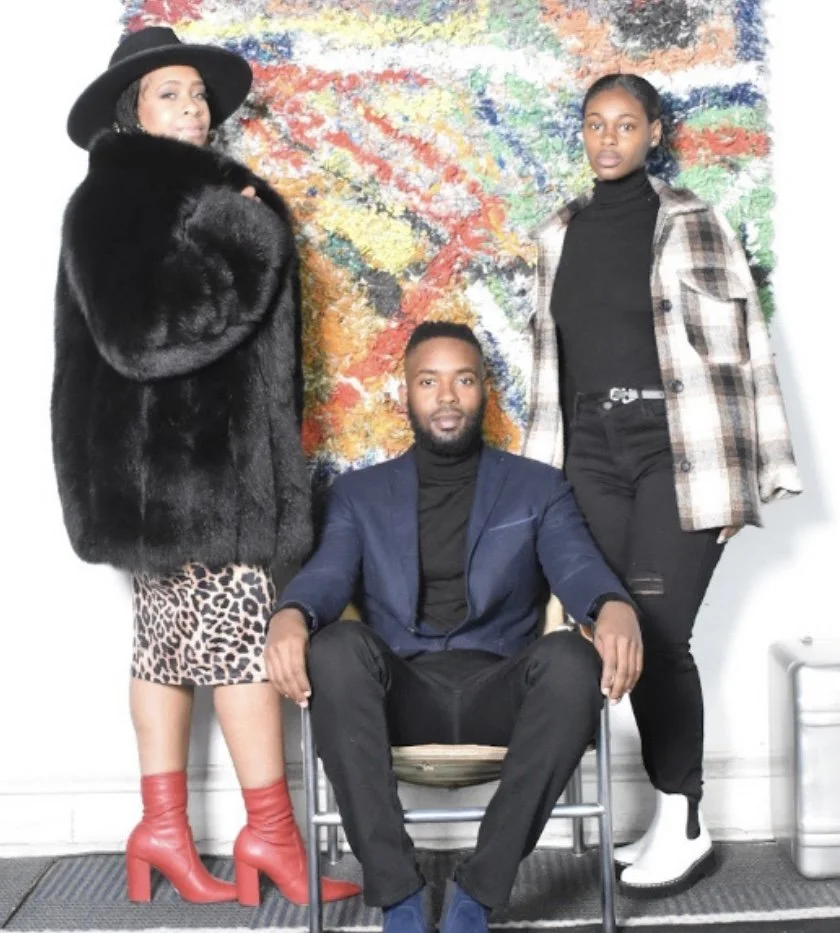 Three people posing in front of a colorful abstract painting. The woman on the left is wearing a black wide-brimmed hat, a black fur coat, leopard print skirt, and red high-heeled boots. The man in the middle is seated, wearing a navy blazer, black turtleneck, and black pants. The woman on the right is standing, dressed in a plaid jacket, black turtleneck, black pants, and white ankle boots.
