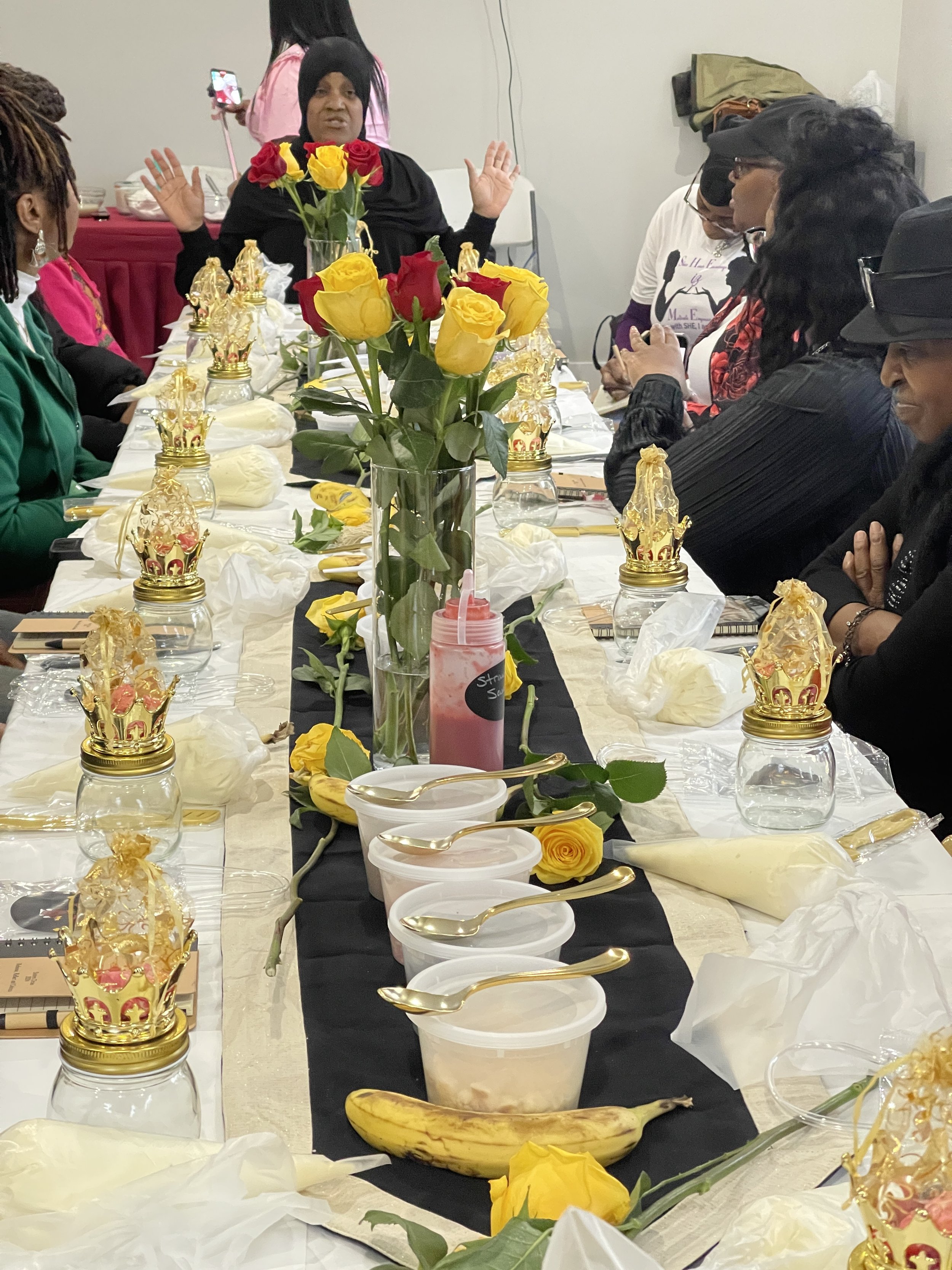 People gathered around a banquet table with yellow roses in a tall glass vase, small jars with gold crown lids, plastic containers with spoons, bananas, a pink smoothie, and wrapped treats. A woman at the head of the table appears to be speaking or making a gesture, with others listening.