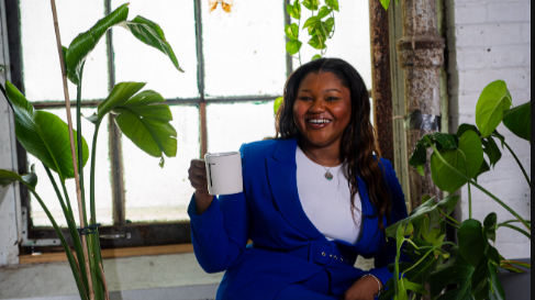A woman holding a white mug while smiling in a bright room with large green plants and a window.