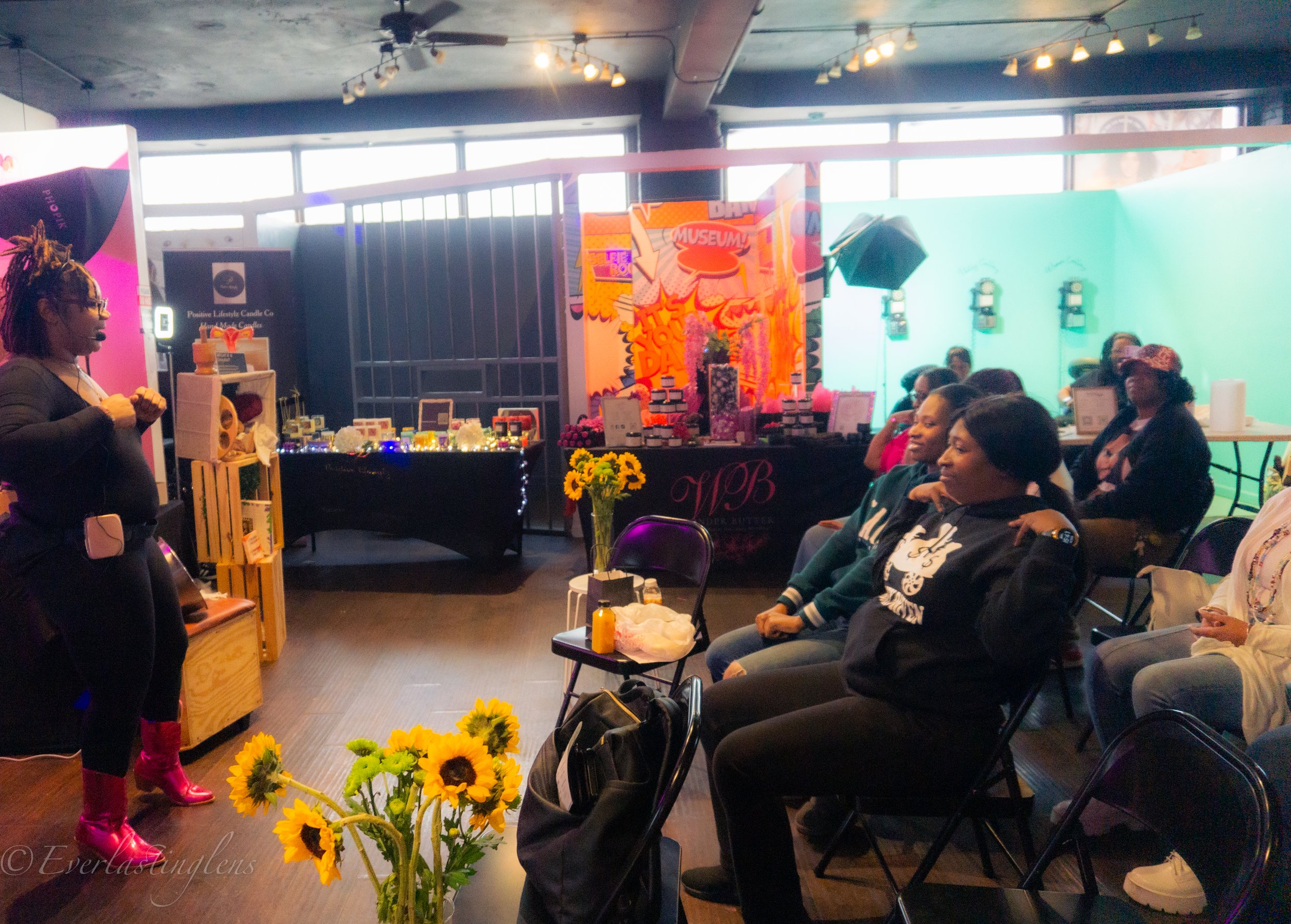 A woman with purple hair and pink boots speaking to an audience of women seated in a room with colorful decor, floral arrangements, and vendor tables.
