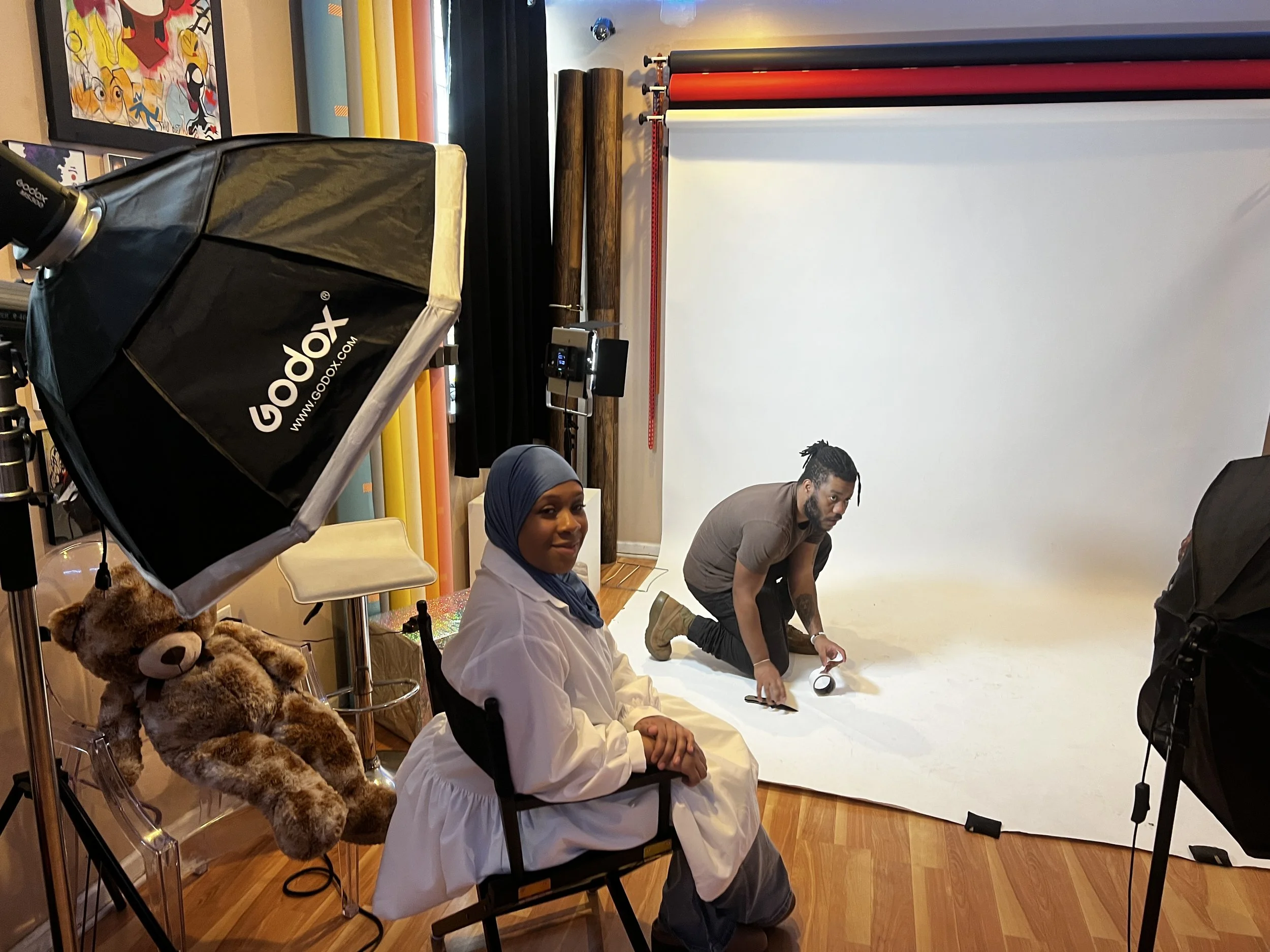 A photoshoot setup with a woman in a white coat and blue headscarf sitting on a chair, a man kneeling on the floor adjusting equipment, a teddy bear on a chair, professional lighting, and a white backdrop.