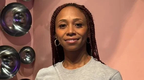 A woman with braided hair and hoop earrings smiling, standing in front of a pink wall with metallic balloons.