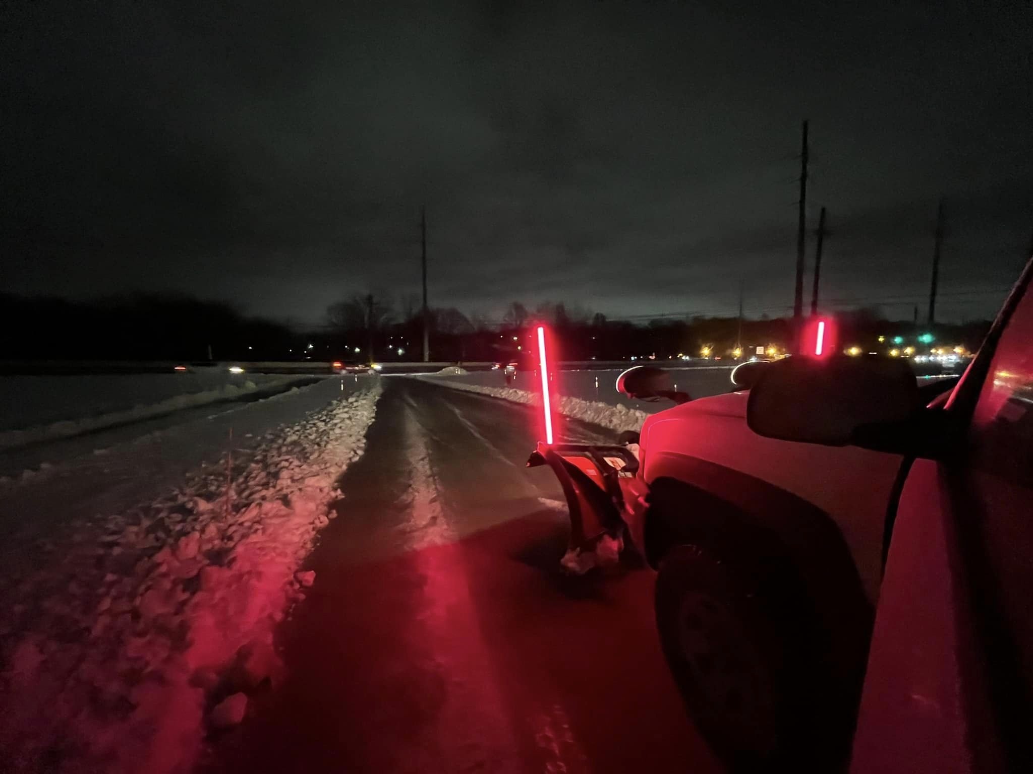 A white pickup truck equipped with a snow plow attachment clearing snow from a road. The truck has a red salt or sand spreader in the back, and snow can be seen on the ground and on the truck.