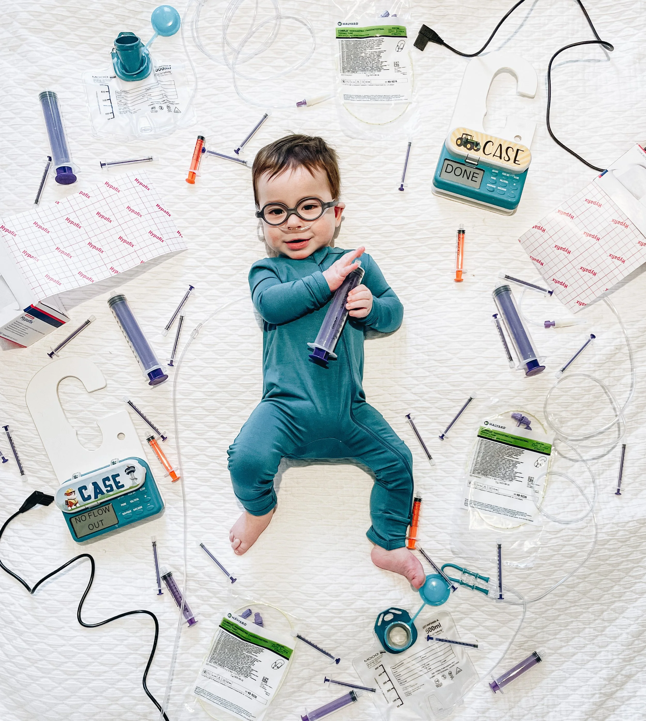 Child lying on a white blanket surrounded by medical supplies, syringes, IV bags, and medical equipment, holding a large syringe, with a playful expression.