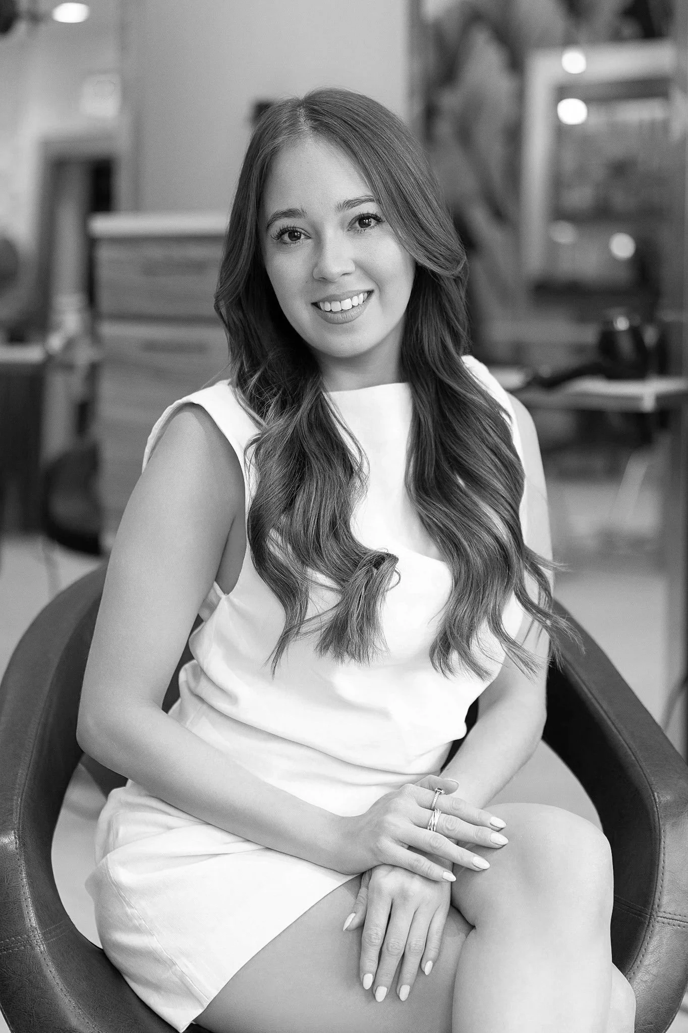 A young woman with long wavy hair smiling, sitting by a window with potted plant on the windowsill, in a well-lit room.