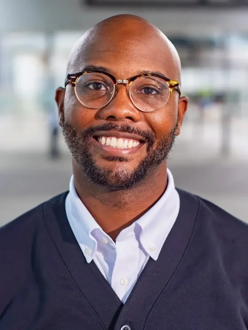 A man wearing glasses, a suit, and a white shirt smiling against a bright blue background.