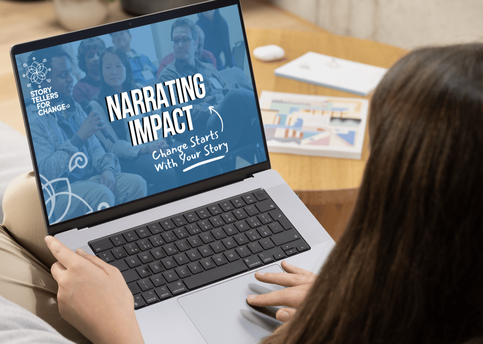 Person sitting at a table using a laptop with a presentation slide on the screen that reads 'Narrating Impact' and 'Change Starts With Your Story,' with a group of people in an audience in the background and documents on the table.