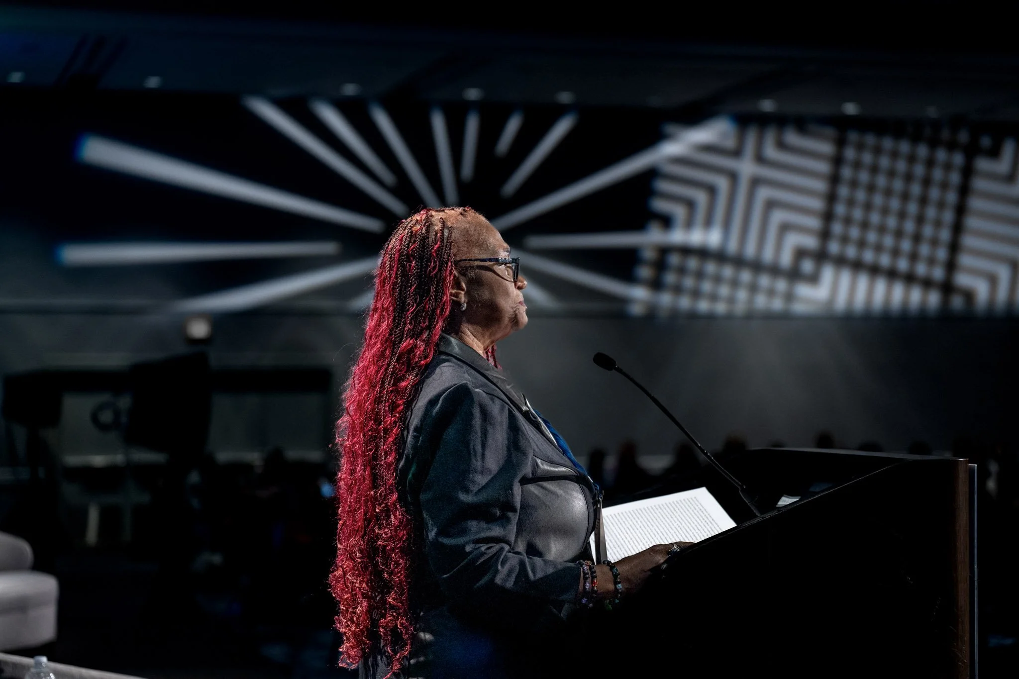 A woman with long braided hair, wearing glasses and a leather jacket, stands at a podium reading from a book in a dark room with modern geometric light patterns on the ceiling.