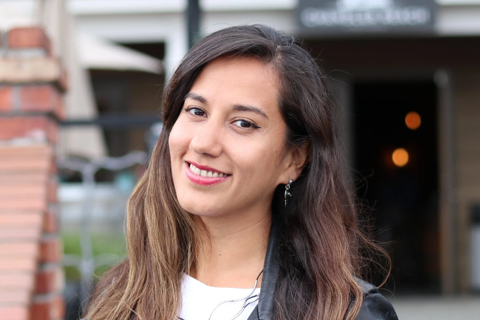 Black and white photo of a smiling woman with long wavy hair, wearing a dark leather jacket and earrings, set against a bright blue background.
