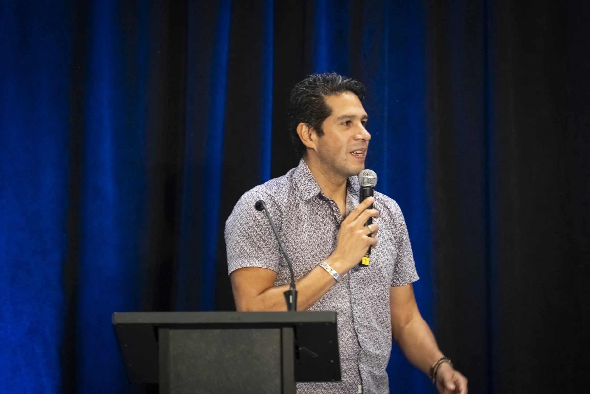 Man with dark, wavy hair speaking into a microphone on stage with blue curtains in the background.