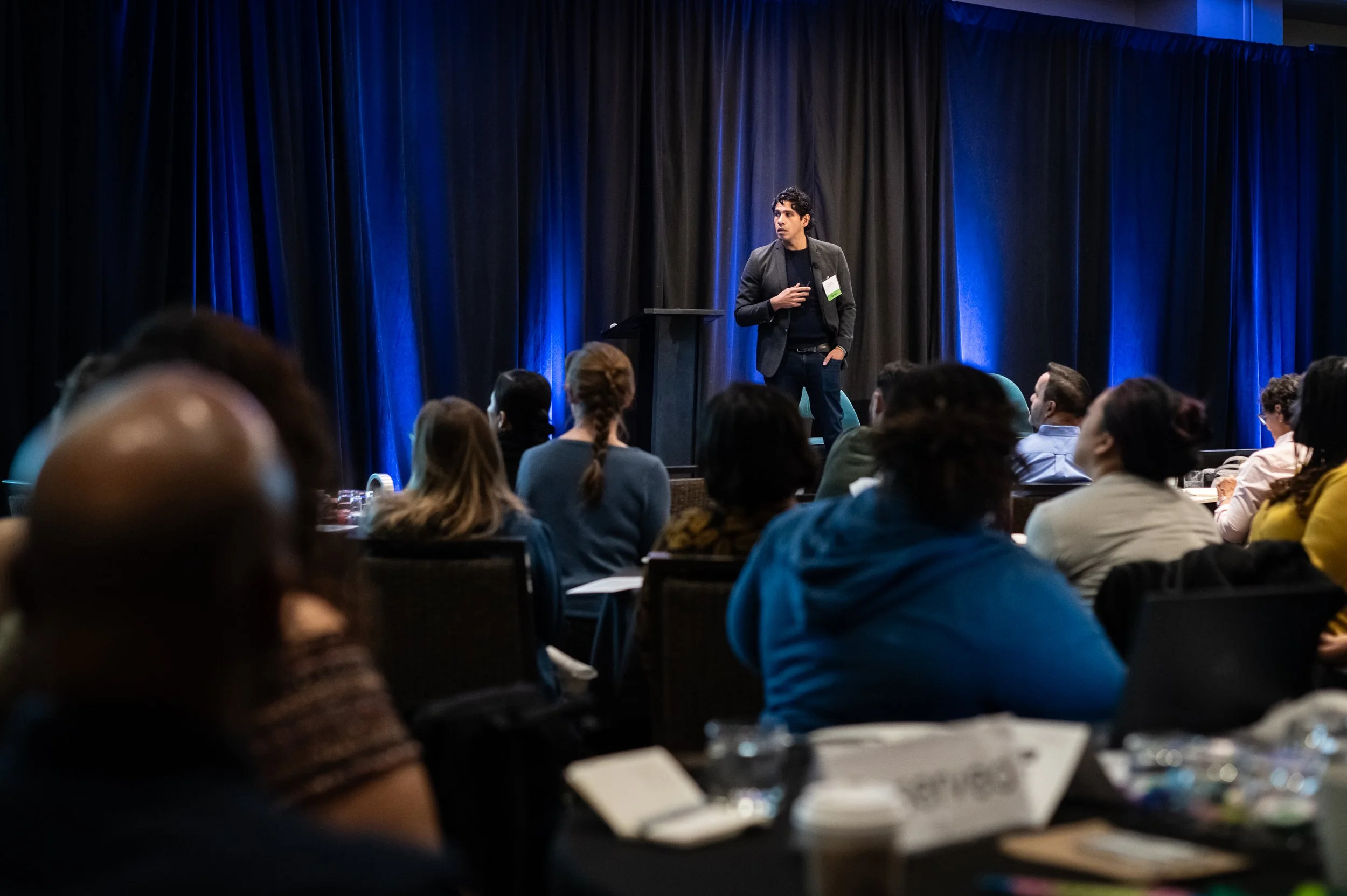 Young woman with braided hair, wearing hoop earrings, speaking into a microphone, smiling, with blue background and orange accent lines near her face.