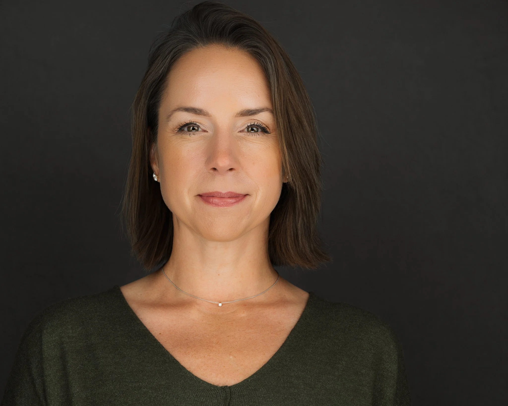 Portrait of a woman with shoulder-length brown hair wearing a dark green top and a delicate necklace, standing against a plain dark background.