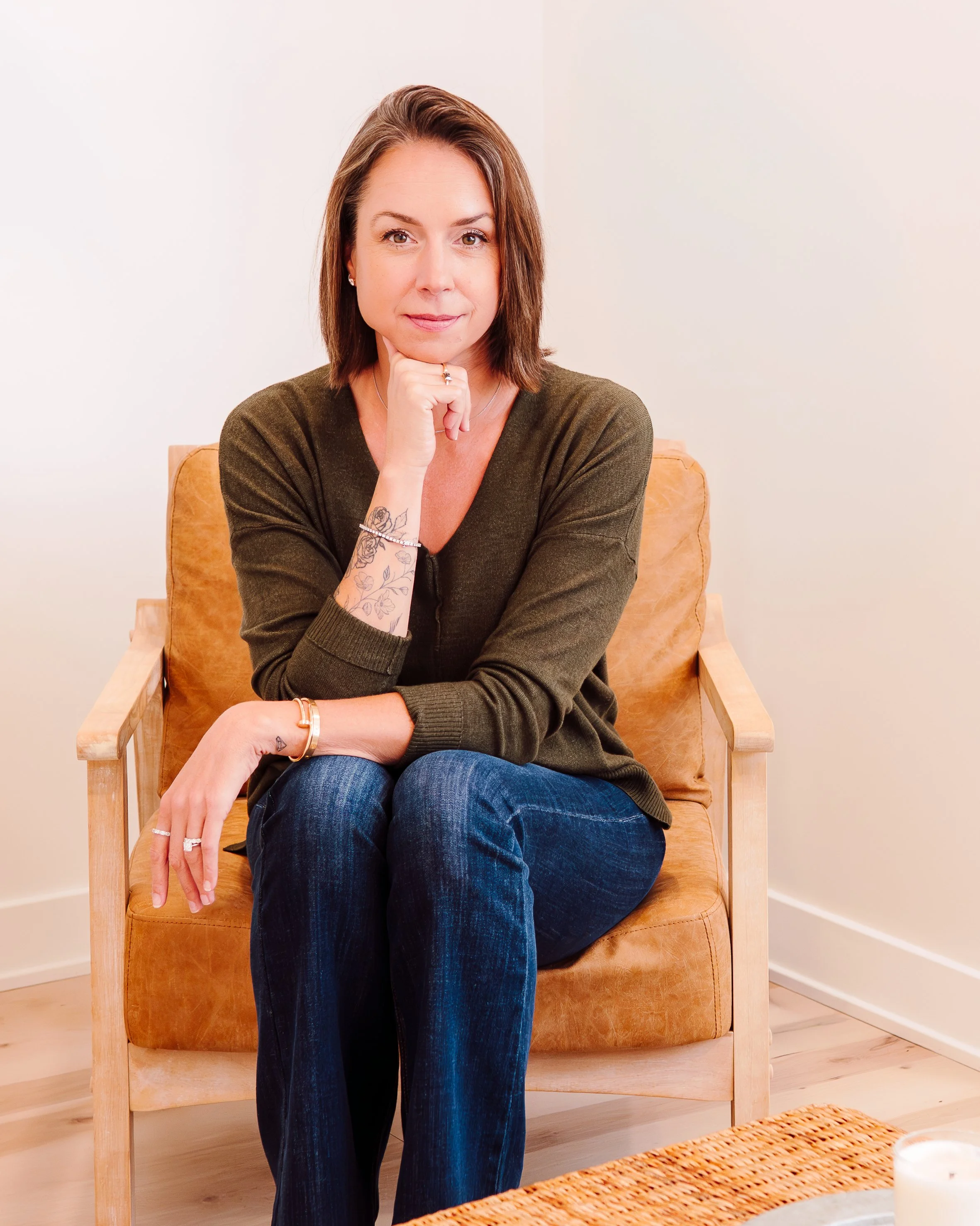 A woman with shoulder-length brown hair, wearing a green sweater and blue jeans, sitting on a tan armchair in a light-colored room, looking directly at the camera with her hand resting on her chin.