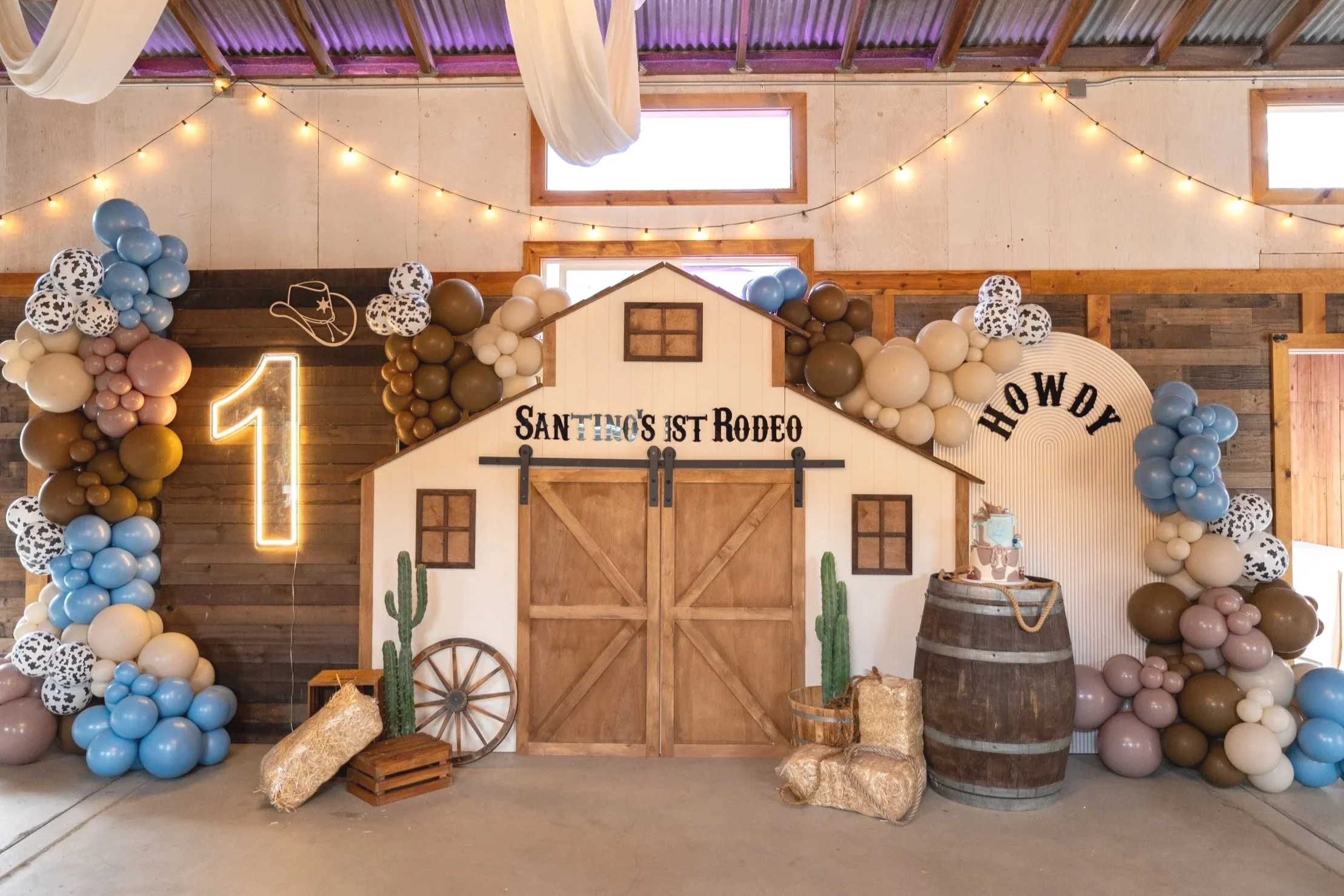 Country-themed birthday decoration with a barn backdrop, balloon arch, large illuminated number 1, cactus decorations, hay bales, a barrel, and a cake on top of the barrel, celebrating a first birthday rodeo theme.