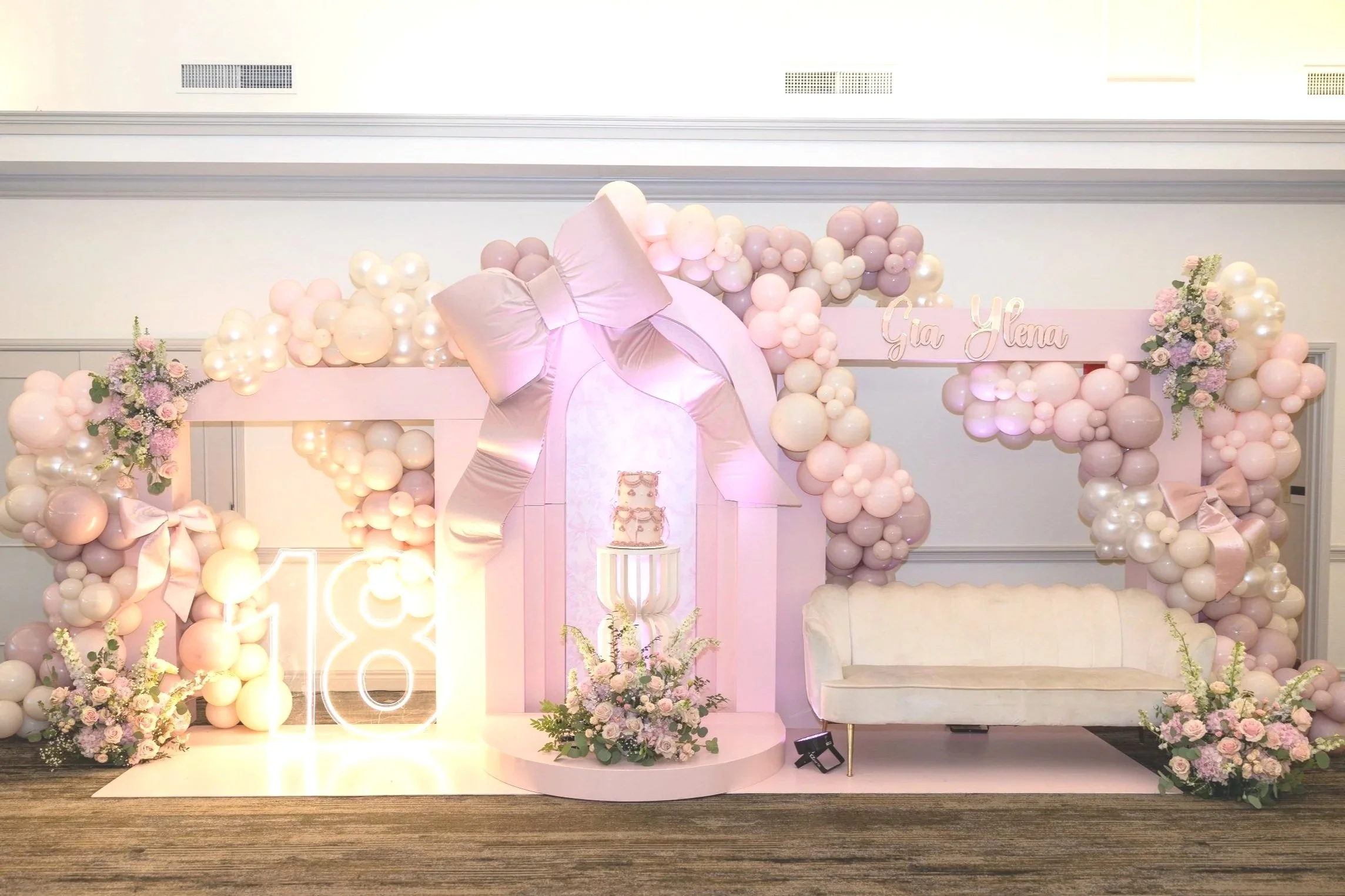 Decorative balloon arch with pink, white, and pastel balloons, flowers, a white sofa, floral arrangements, and a cake display with the name Gia Helena in a pastel-colored event setting.