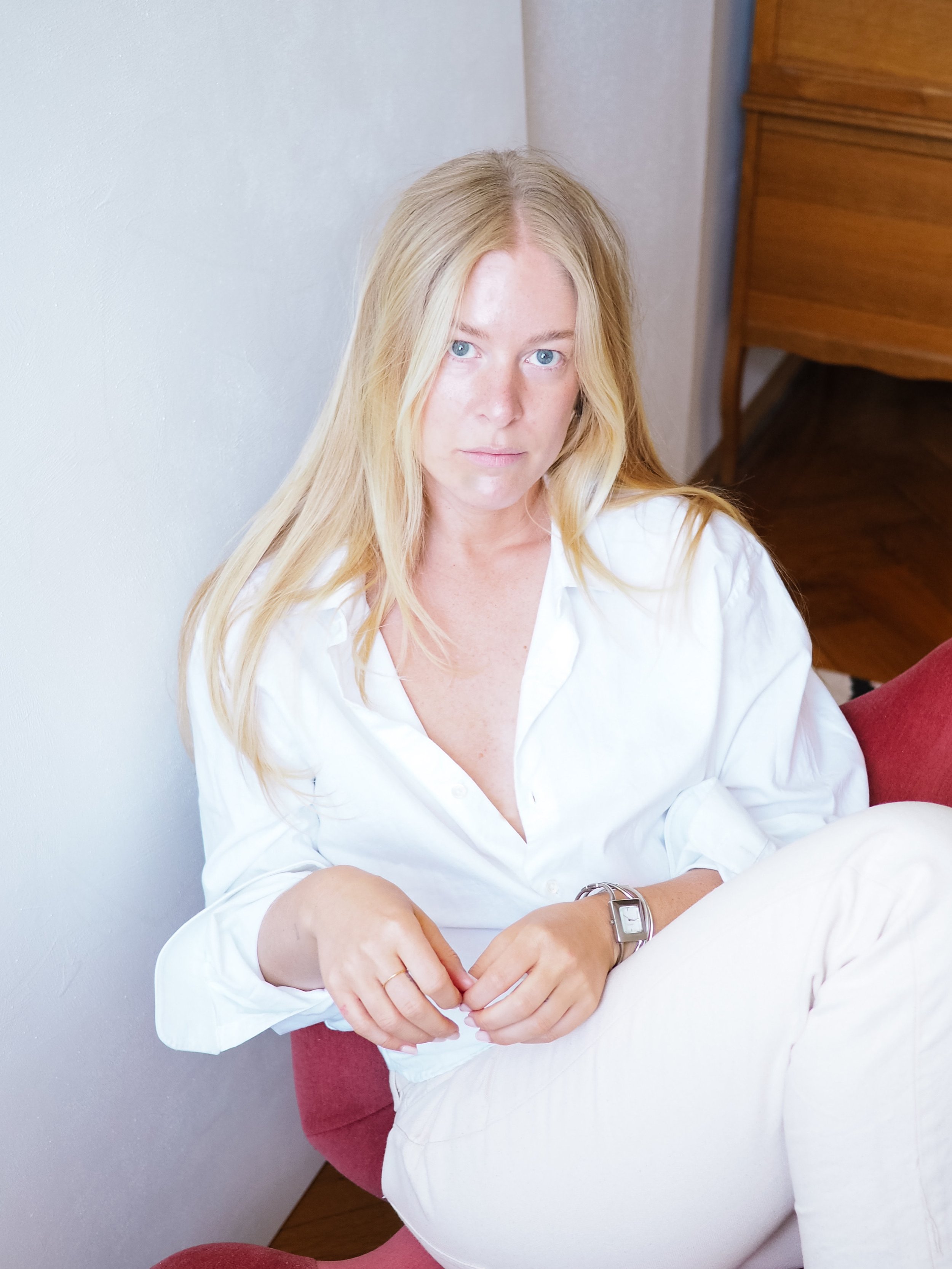 A woman with long blonde hair and blue eyes sitting on a red armchair, wearing a white shirt, looking directly at the camera.