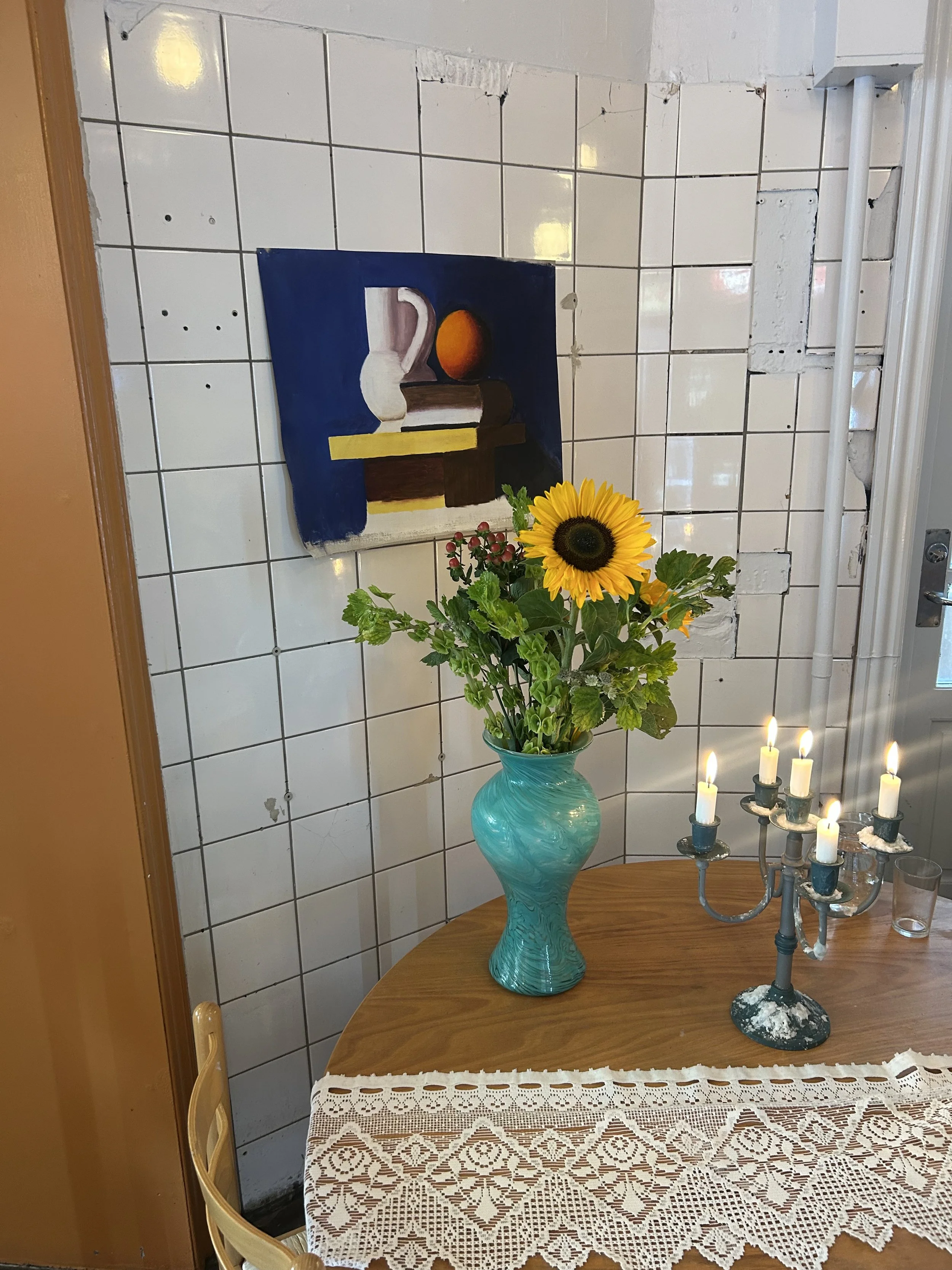 A wooden table with a white lace tablecloth, a turquoise vase with a sunflower and other green and red plants, a candelabra with lit candles, and a framed still life painting of a white pitcher and an orange on dark blue background on a tiled white wall in the background.