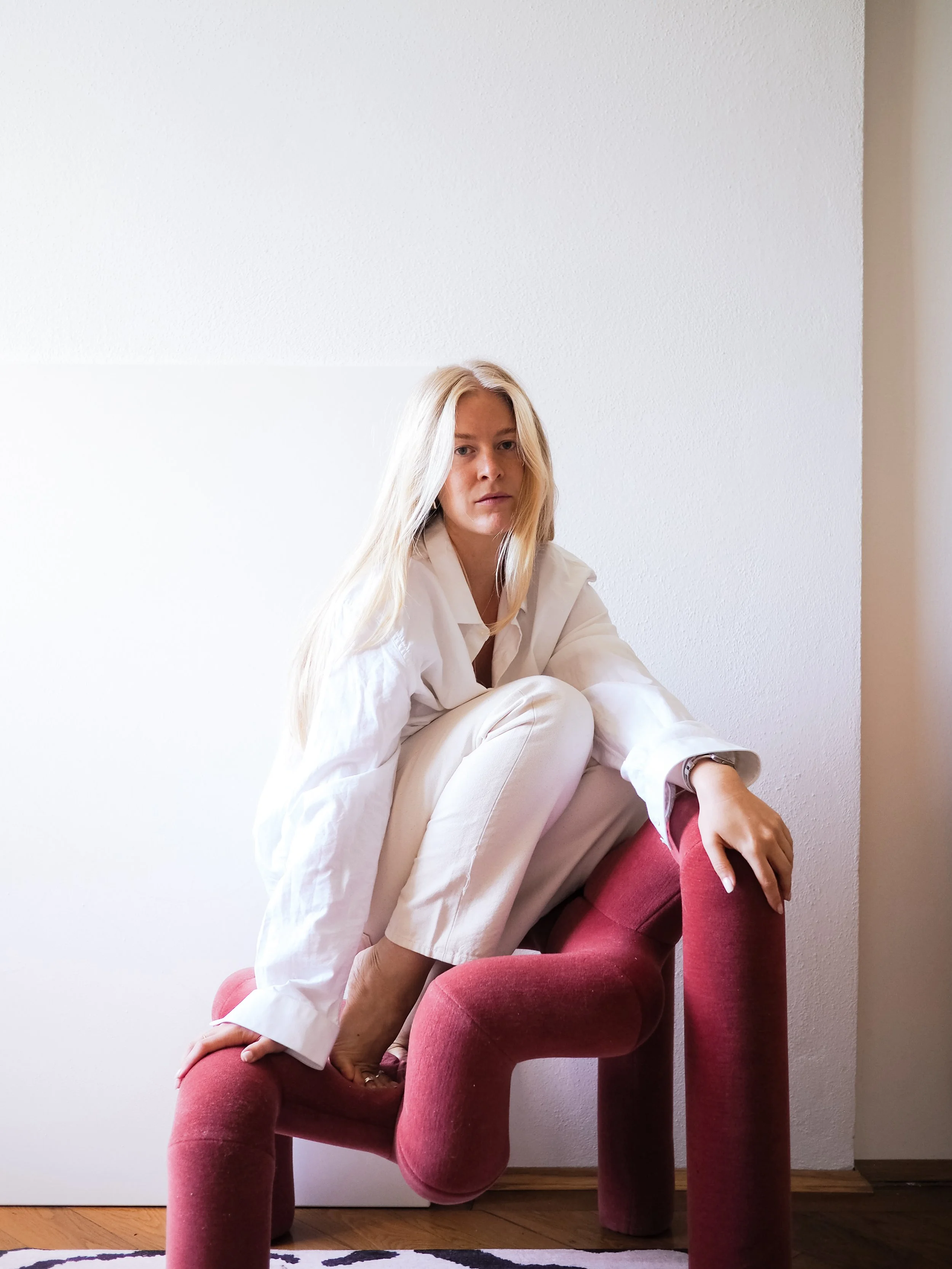 A woman with blonde hair wearing a white shirt and red pants sitting on a pink velvet chair in front of a plain white wall.