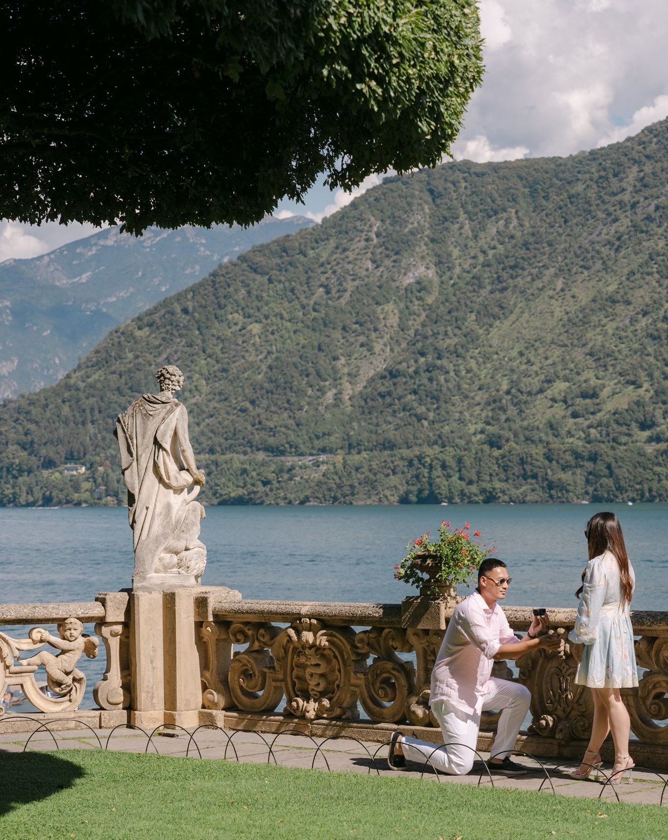 A Lake Como proposal at Villa del Balbianello, early afternoon, on the terrace above the water.

Lily and Jerry. A stone balustrade, a statue with its back to the lake, red geraniums in the urn. One knee on the flagstones, a ring box opening, and her