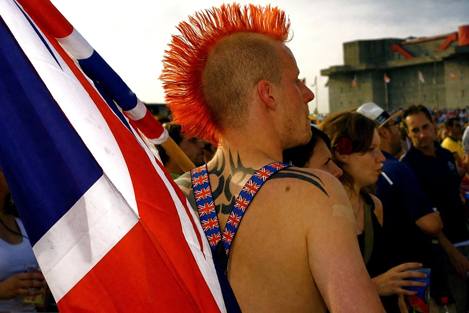 A person with a Mohawk hairstyle dyed orange, standing in a crowd during a celebration or event. The person is shirtless, has tattoos on their shoulder and neck, and is holding a large British flag while wearing a suspenders with Union Jack pattern. Others around are in casual attire, some holding drinks, with a large building in the background.