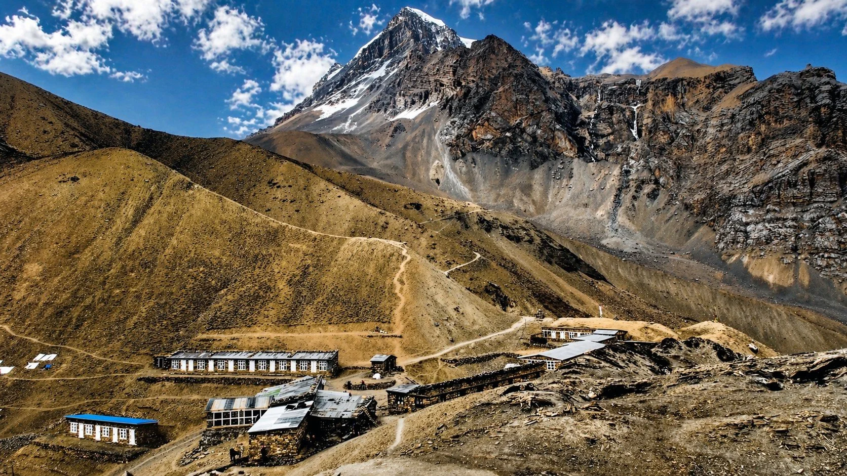 Mountain landscape with a snow-capped peak in the background, rugged slopes, and scattered buildings at the base, under a partly cloudy sky.