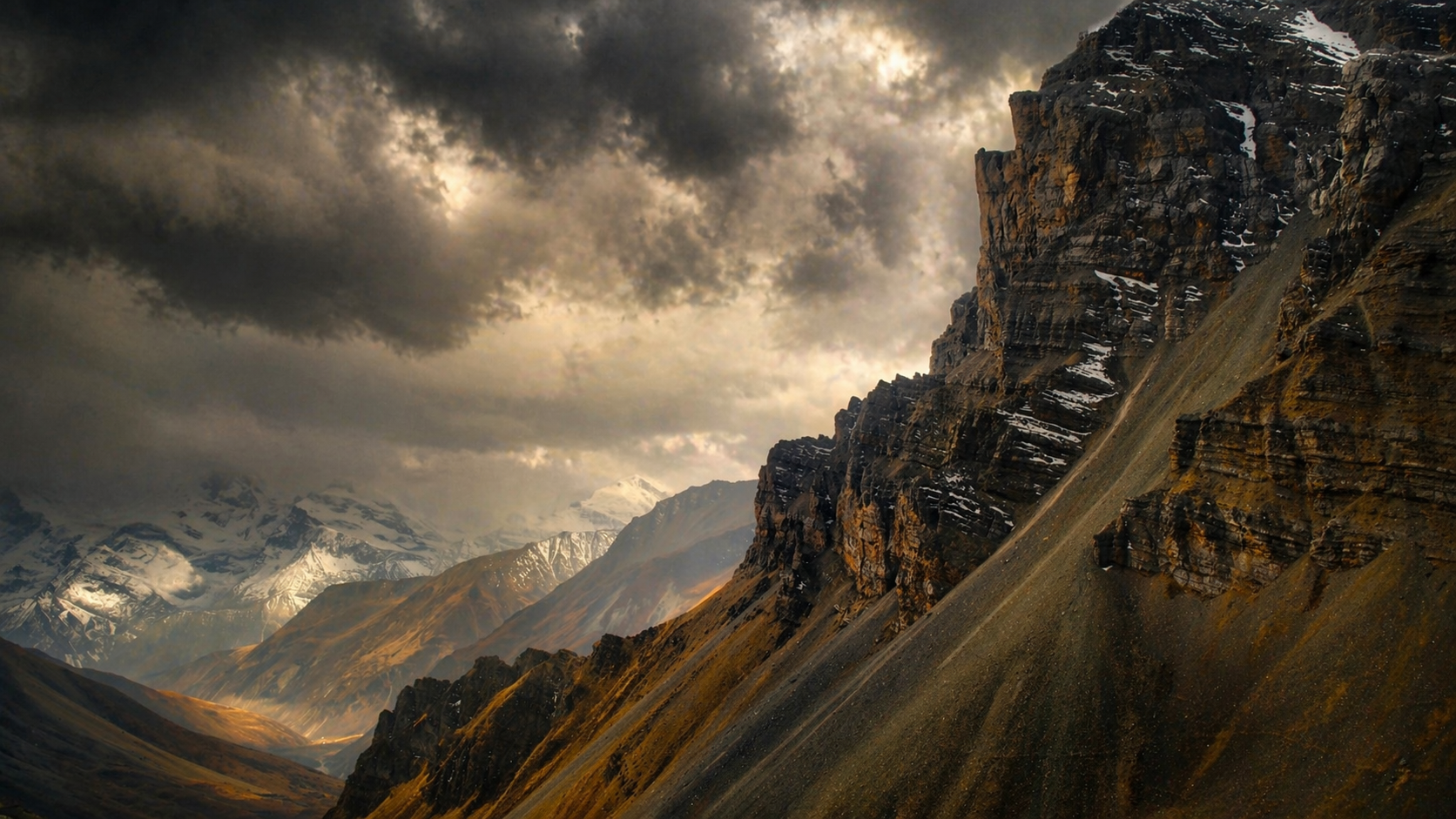 A rugged mountain landscape with dark, cloudy skies, steep rocky mountain slopes with patches of snow, and distant snow-capped peaks under dramatic weather conditions.