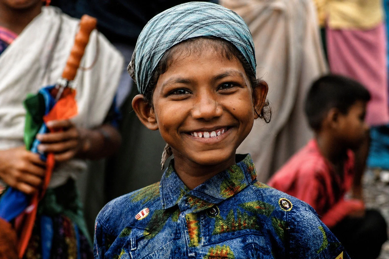 A smiling young girl with a gap-tooth smile, wearing a blue patterned shirt and a headscarf, standing among others in a crowded outdoor setting.