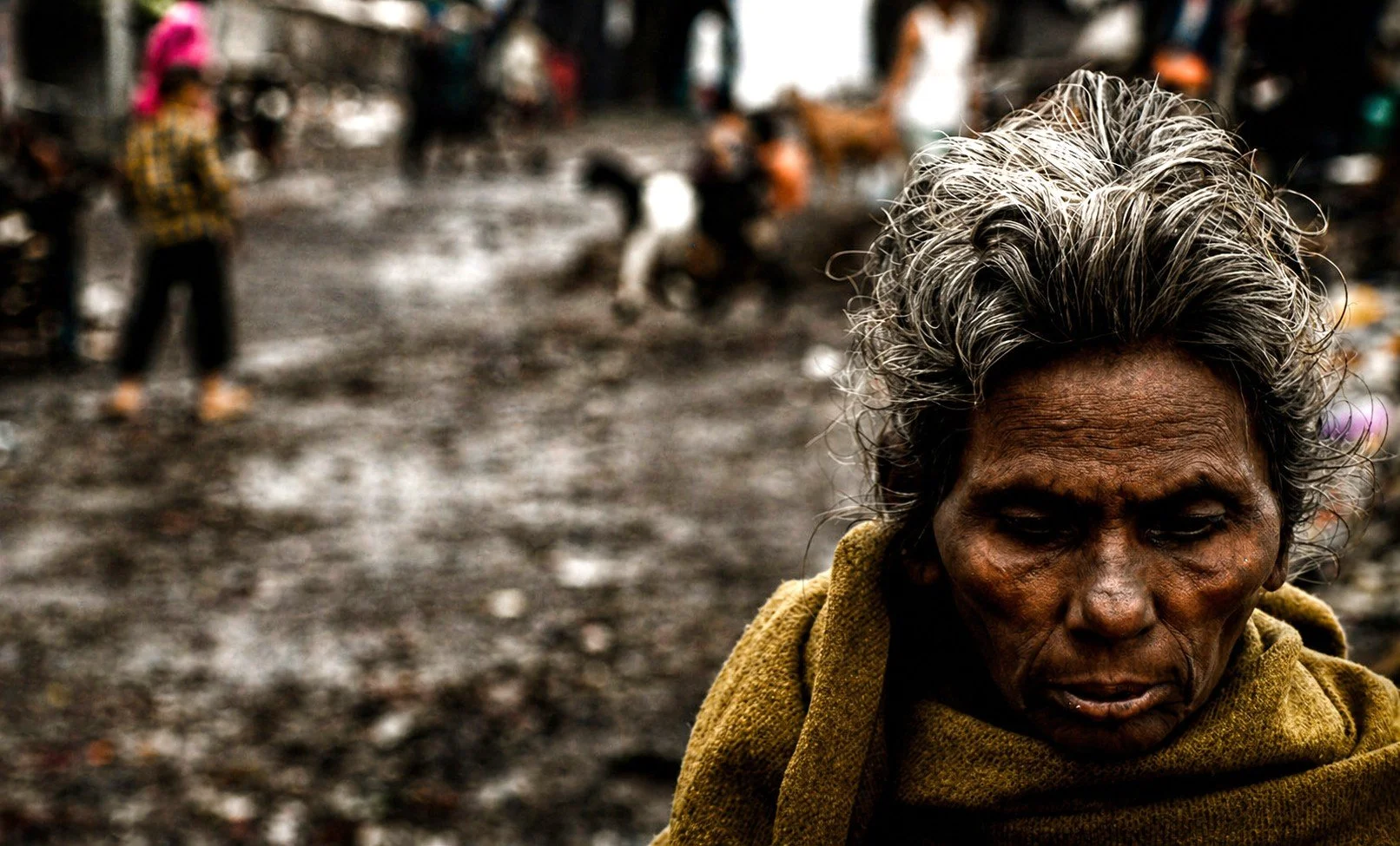 Close-up of an elderly woman with gray hair and deep wrinkles, wearing a yellow scarf, standing in a muddy area with blurred people and structures in background.