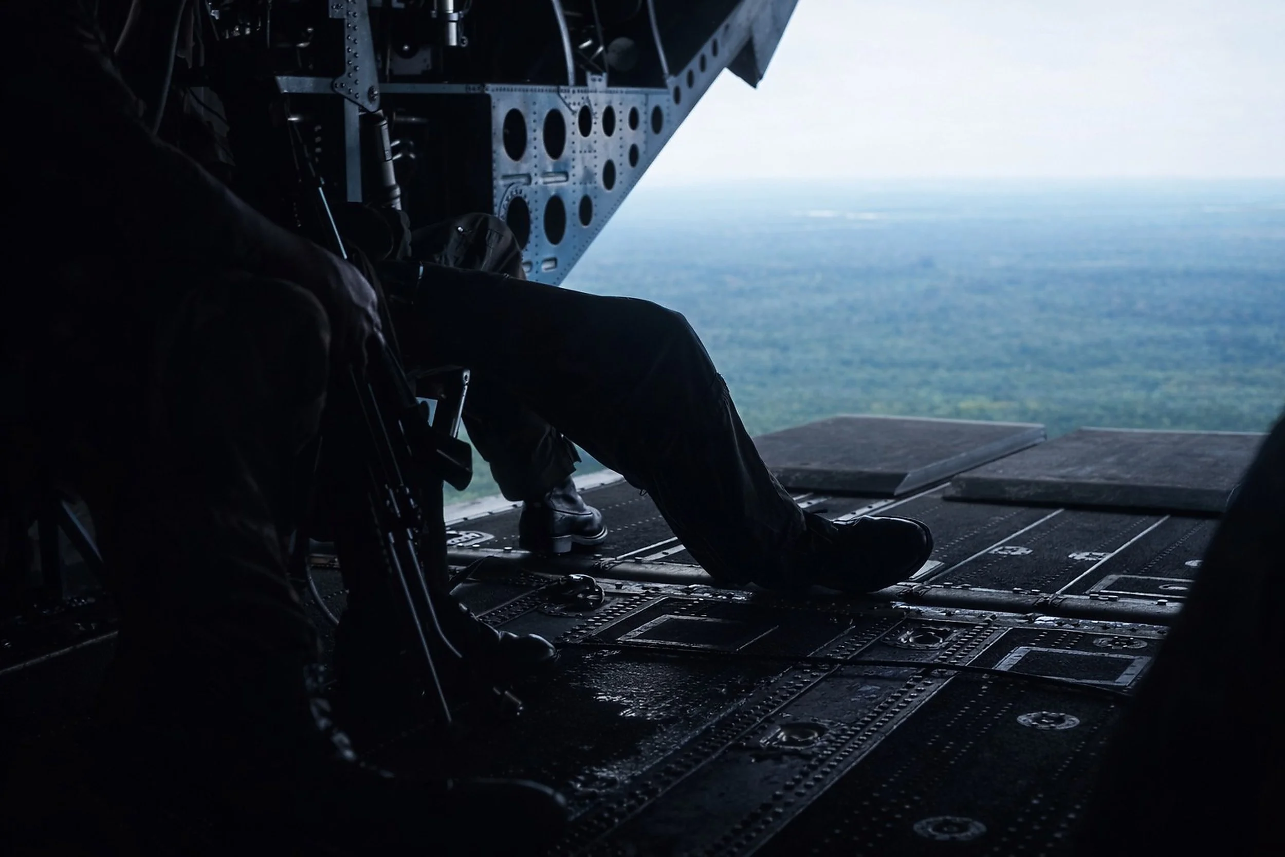 A person sitting on the open rear ramp of an aircraft, looking out over the landscape of forests and sky.