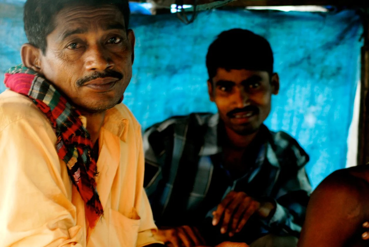 Two men sitting in front of a blue fabric backdrop, one in a light-colored shirt with a red and green checkered cloth around his neck, and the other in a dark plaid shirt, both looking at the camera.