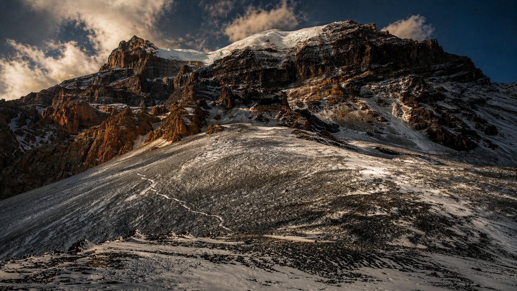 Snow-covered mountain with rugged rocky cliffs under a partly cloudy sky during sunset or sunrise.