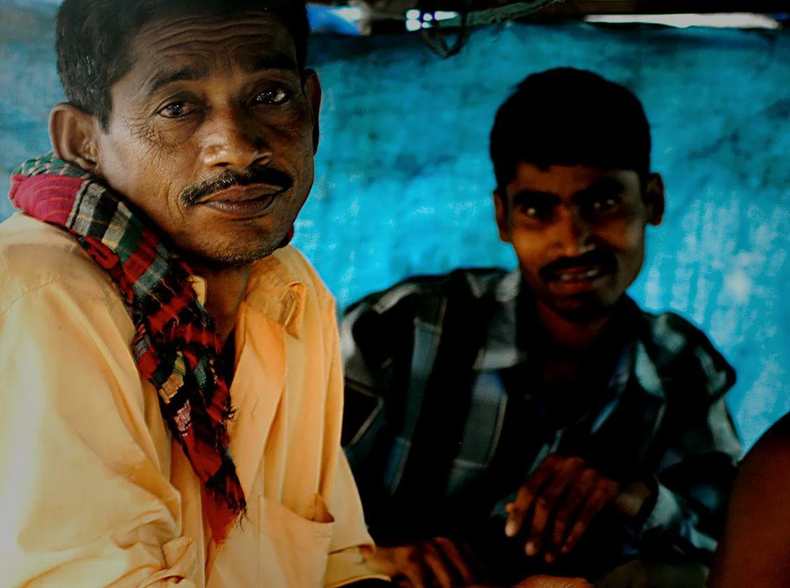 Two men sitting indoors, one older with a mustache, wearing a beige shirt and a red plaid scarf, and a younger man in a checkered shirt, against a blue background.