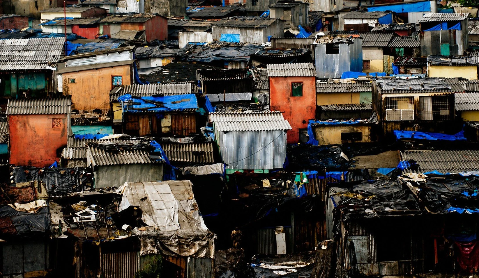 A densely packed slum area with numerous small, makeshift houses made of corrugated metal and various materials, with some roofs covered in blue tarps, in an urban environment.
