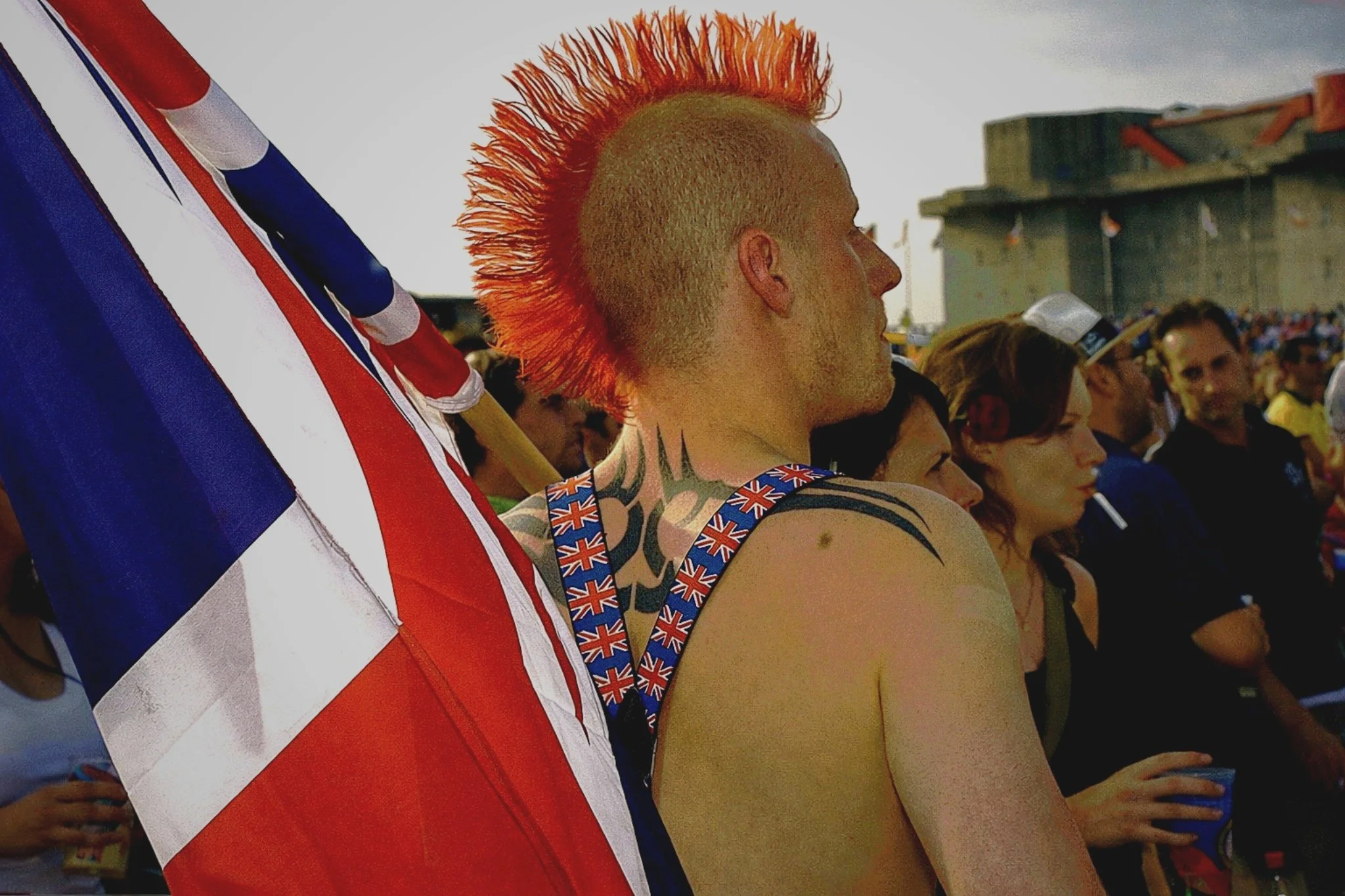 A man with a bright orange mohawk hairstyle, tattoos, and wearing suspenders decorated with Union Jack flags stands among a crowd at an outdoor event. The American flag is draped over his shoulder.