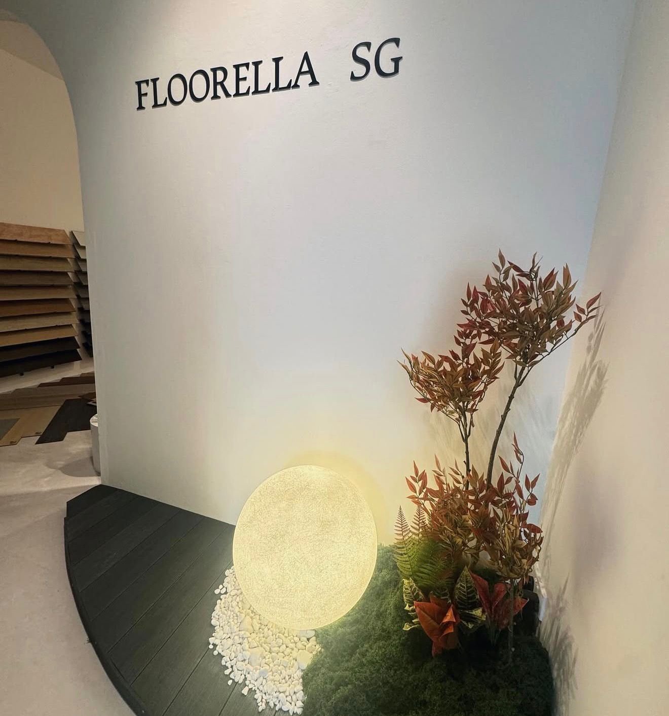 Interior corner with a white wall that has black letters spelling 'FLOORELLA SG', a decorative illuminated sphere, brown and green plants, and a patch of green moss or grass on the floor.