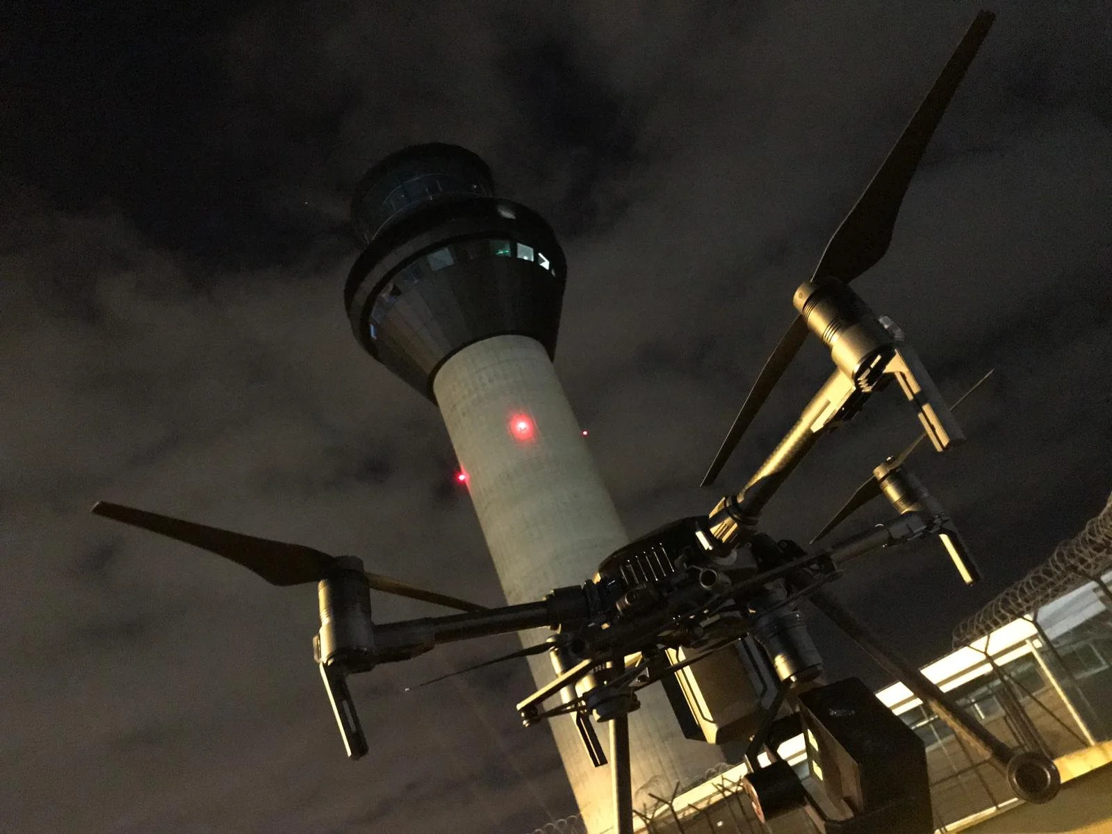 Nighttime view of an airport control tower with a drone or robotic device in the foreground, against a cloudy sky.