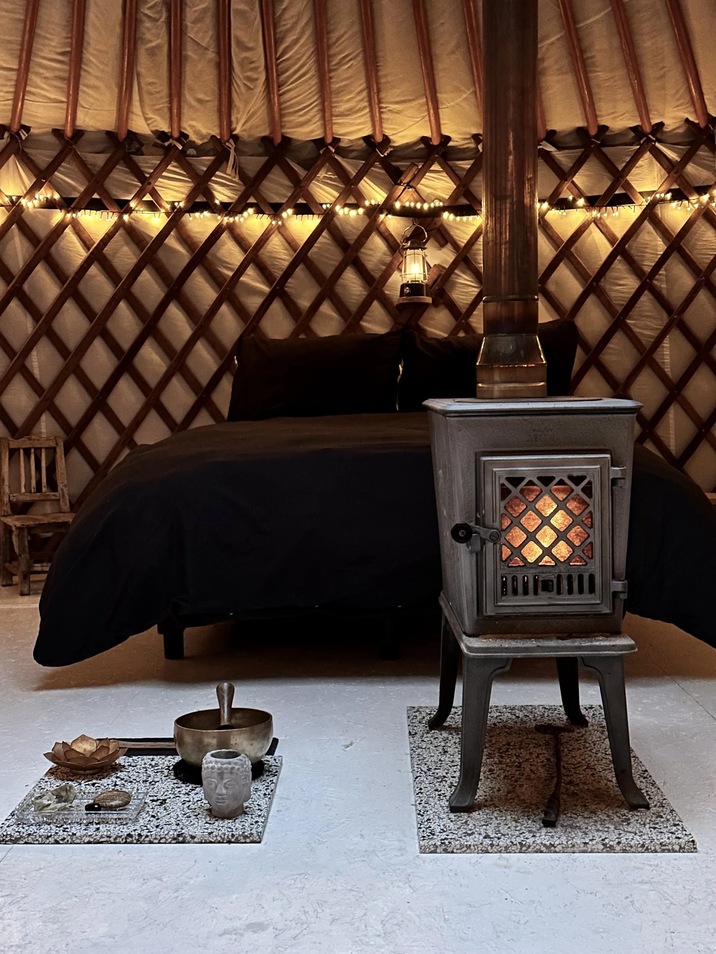 Interieur van een yurt met een bed, een houtkachel, en decoratieve objecten zoals een klankschaal, een beeldje, en een kleine stoel, met warme verlichting en lichte romige vloer.