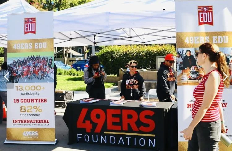 Two women standing behind a table under a red canopy at a fire safety booth, with fire department and fire truck graphics on display and a sign that says Santa Clara Fire Department.