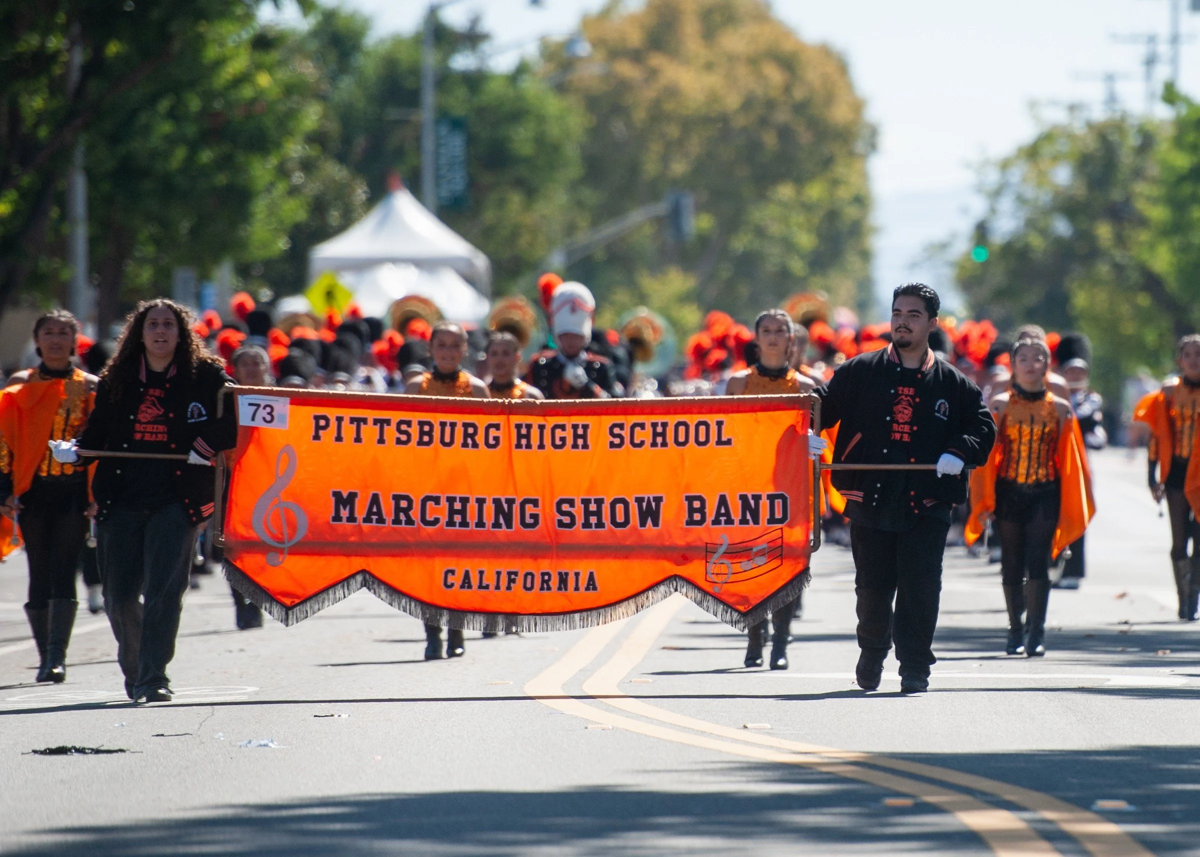 A parade on a sunny day featuring a marching band dressed in blue and white uniforms, with two girls holding a blue banner with Chinese characters and English text that reads 'Falun Dafa.' The crowd is watching from the sides, with trees and utility poles in the background.