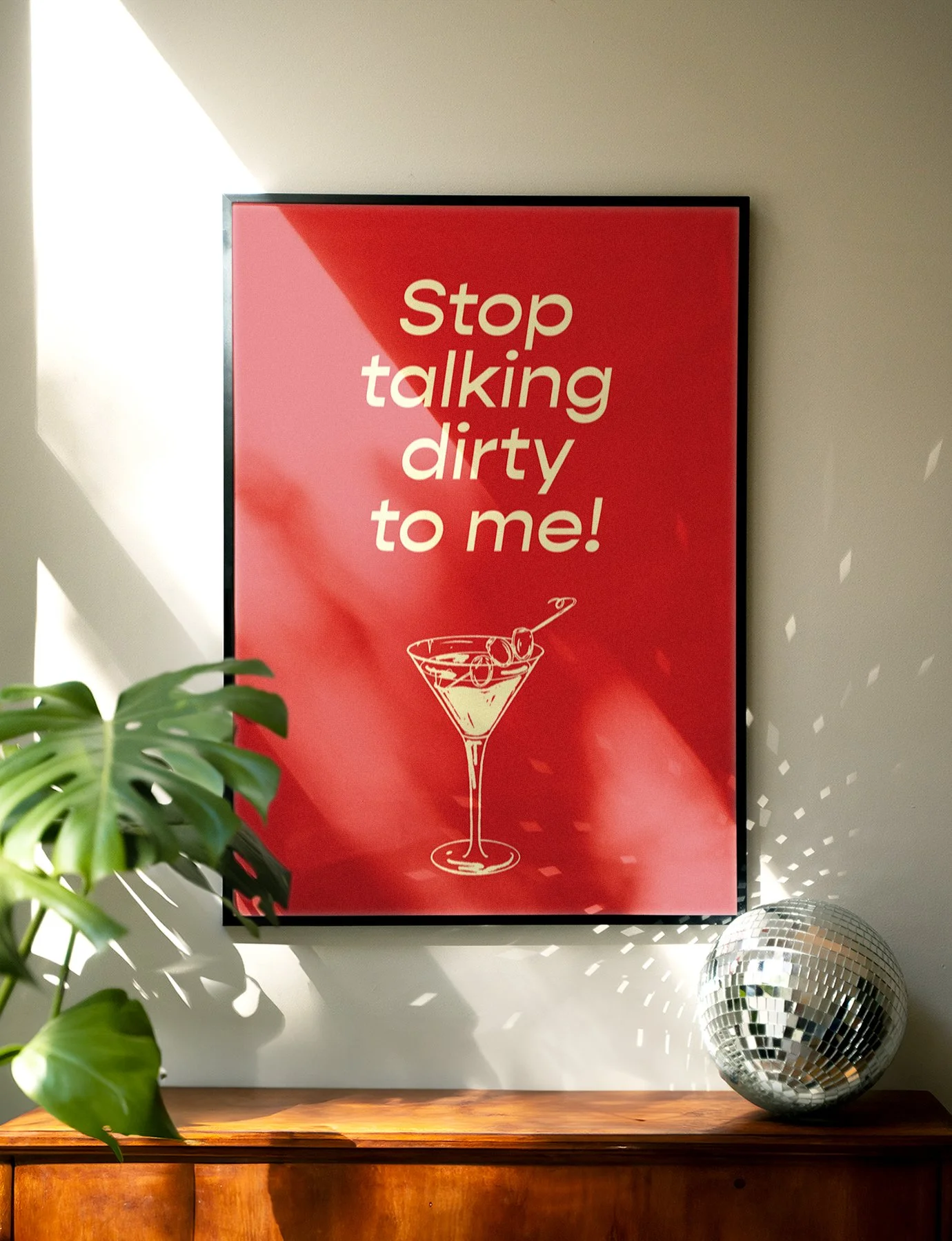 Red framed wall decoration with the phrase 'Stop talking dirty to me!' in white text, a drawing of a cocktail glass with olives, next to a potted plant and a disco ball reflected in direct light.