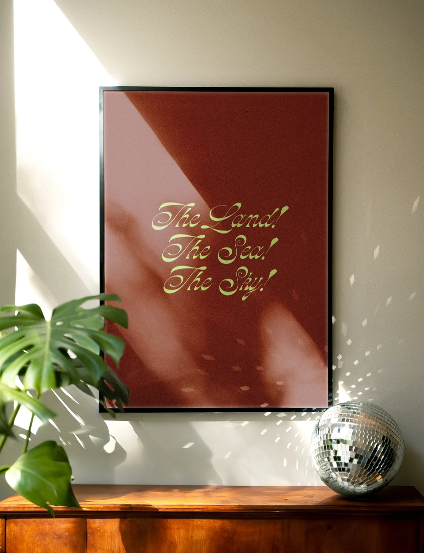 A framed quote on a wall reading "The Land! The Sea! The Sky!" with a green houseplant in the foreground and a small disco ball on a wooden surface beneath.