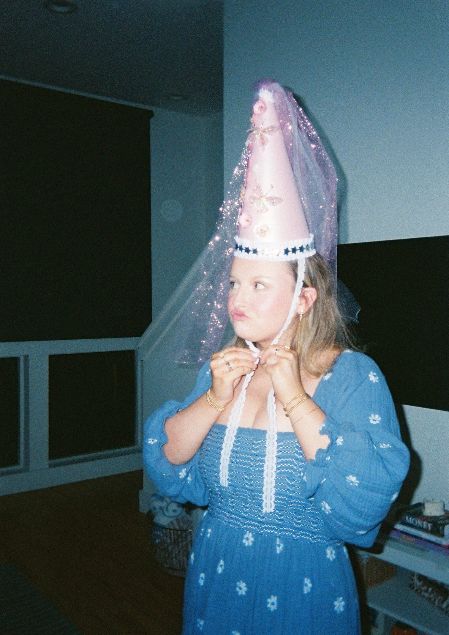 Woman adjusting a pink cone-shaped birthday hat with purple tulle and butterfly decorations, wearing a blue dress with white embroidered flowers.