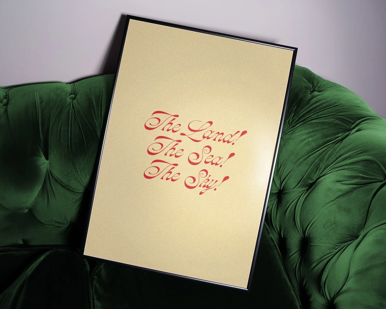 A framed quote resting on a green velvet tufted sofa. The quote says, "The Land! The Sea! The Sky!" in red cursive font.