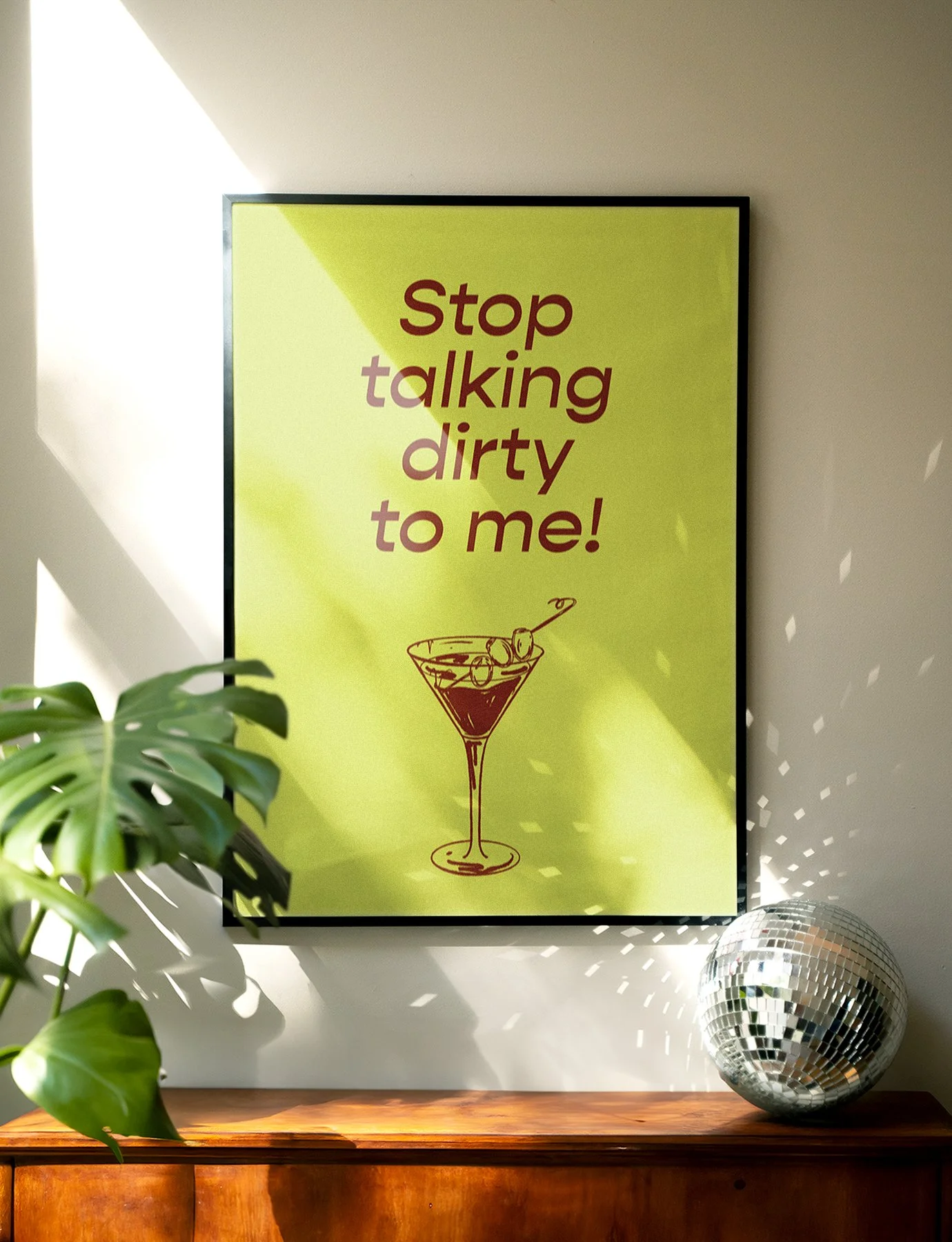 A yellow poster with the message "Stop talking dirty to me!" written in burgundy letters, featuring a simple illustration of a cocktail glass with olives and a cherry, hanging on a wall next to a green plant and a disco ball on a wooden surface.