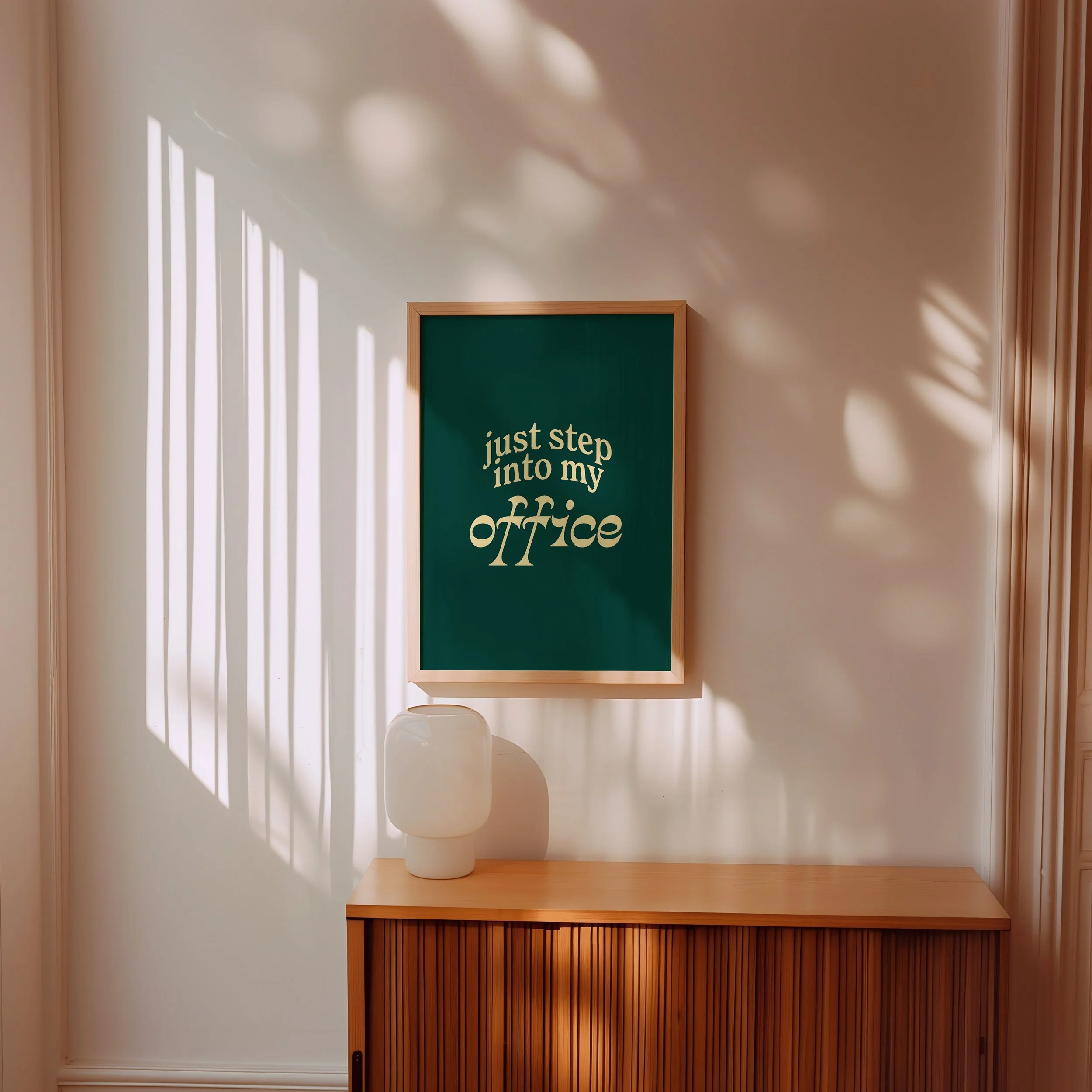 A corner of a room with a green framed chalkboard reading 'just step into my office'. The room features a wooden desk, a white decorative jar, and a wooden sliding door. Sunlight creates striped shadow patterns on the wall.