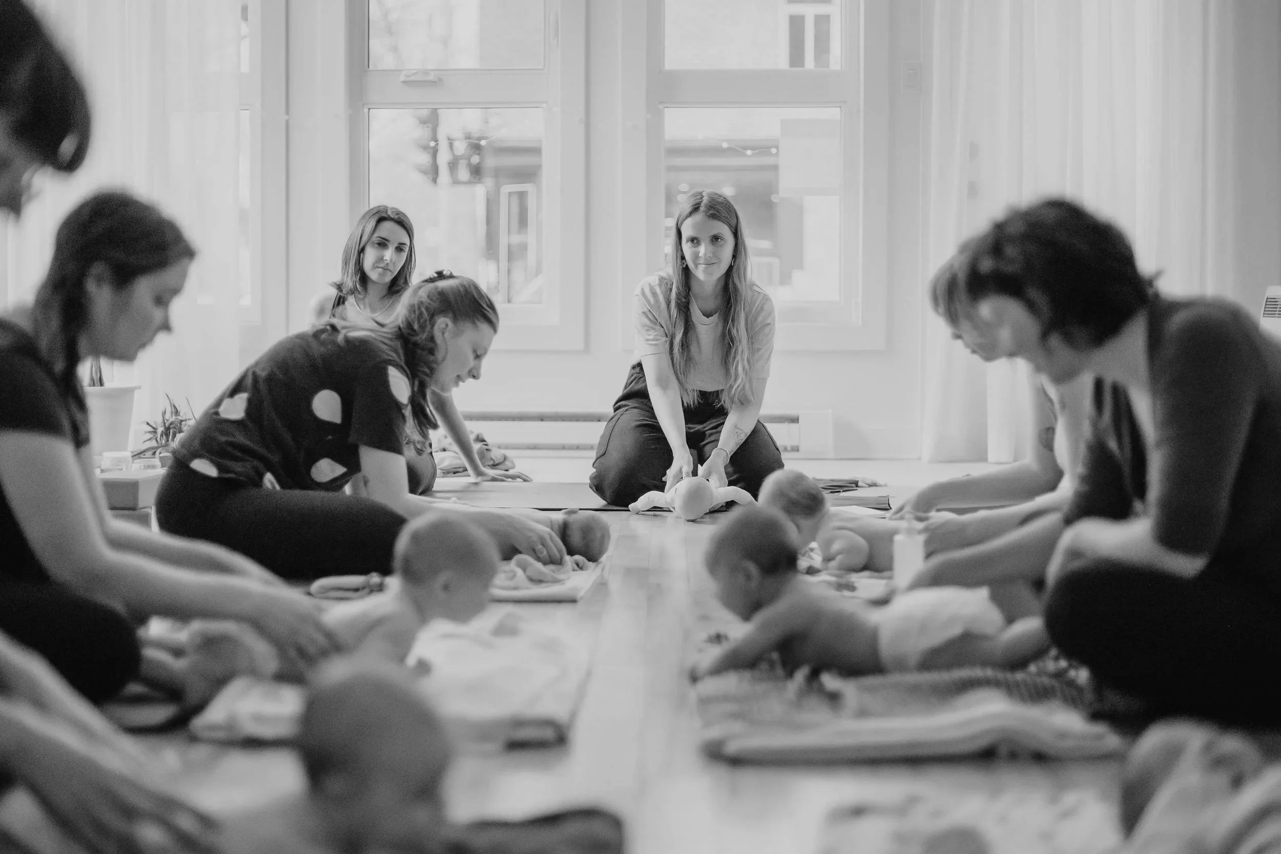 Groupe de femmes assises par terre, participant à un atelier ou une séance de groupe, avec une femme assise au centre en train d'expliquer ou de présenter quelque chose, dans une pièce lumineuse avec de grandes fenêtres.