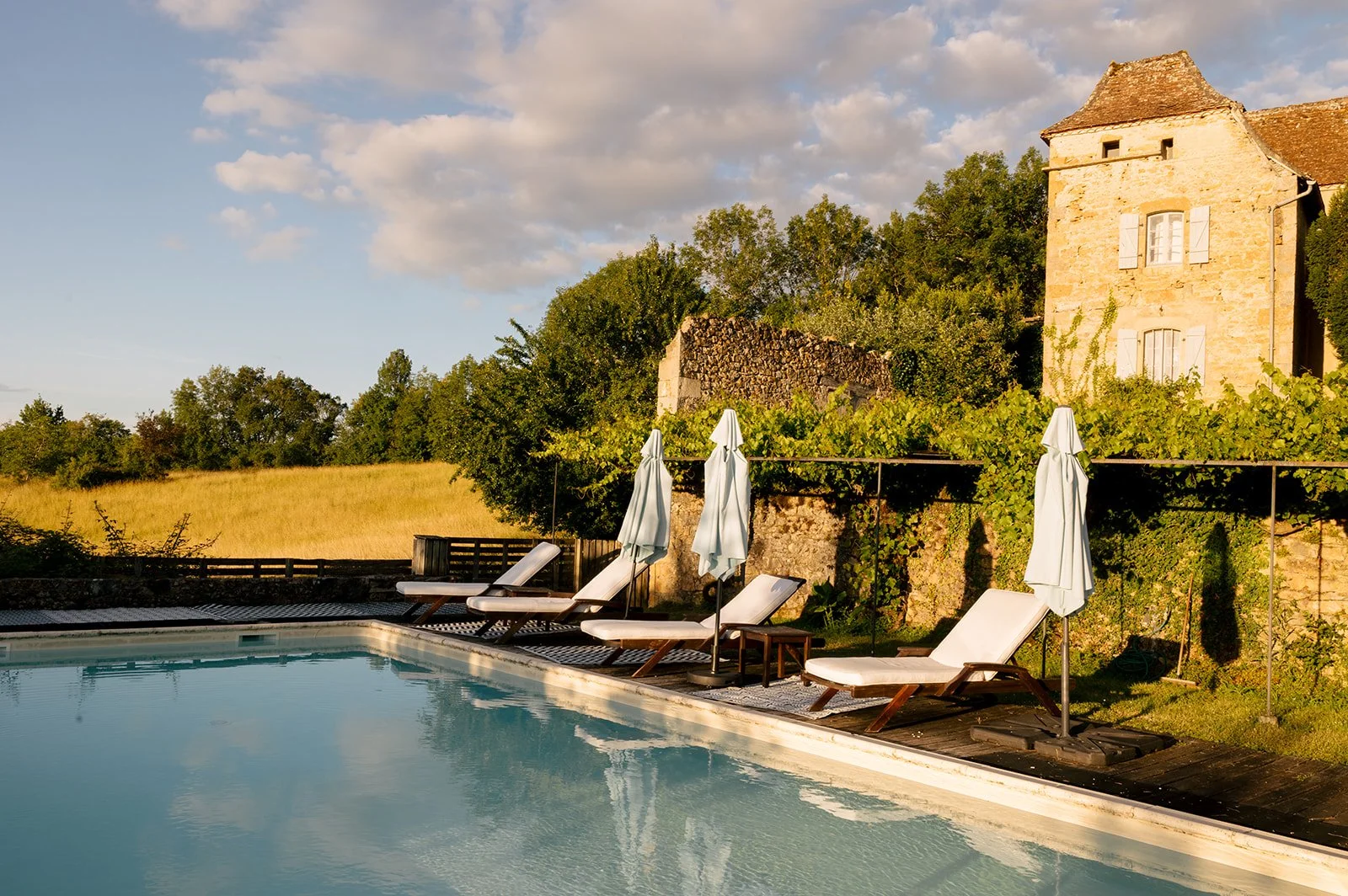 A swimming pool with lounge chairs and umbrellas on a patio, with an old stone house, trees, and a field in the background under a partly cloudy sky.