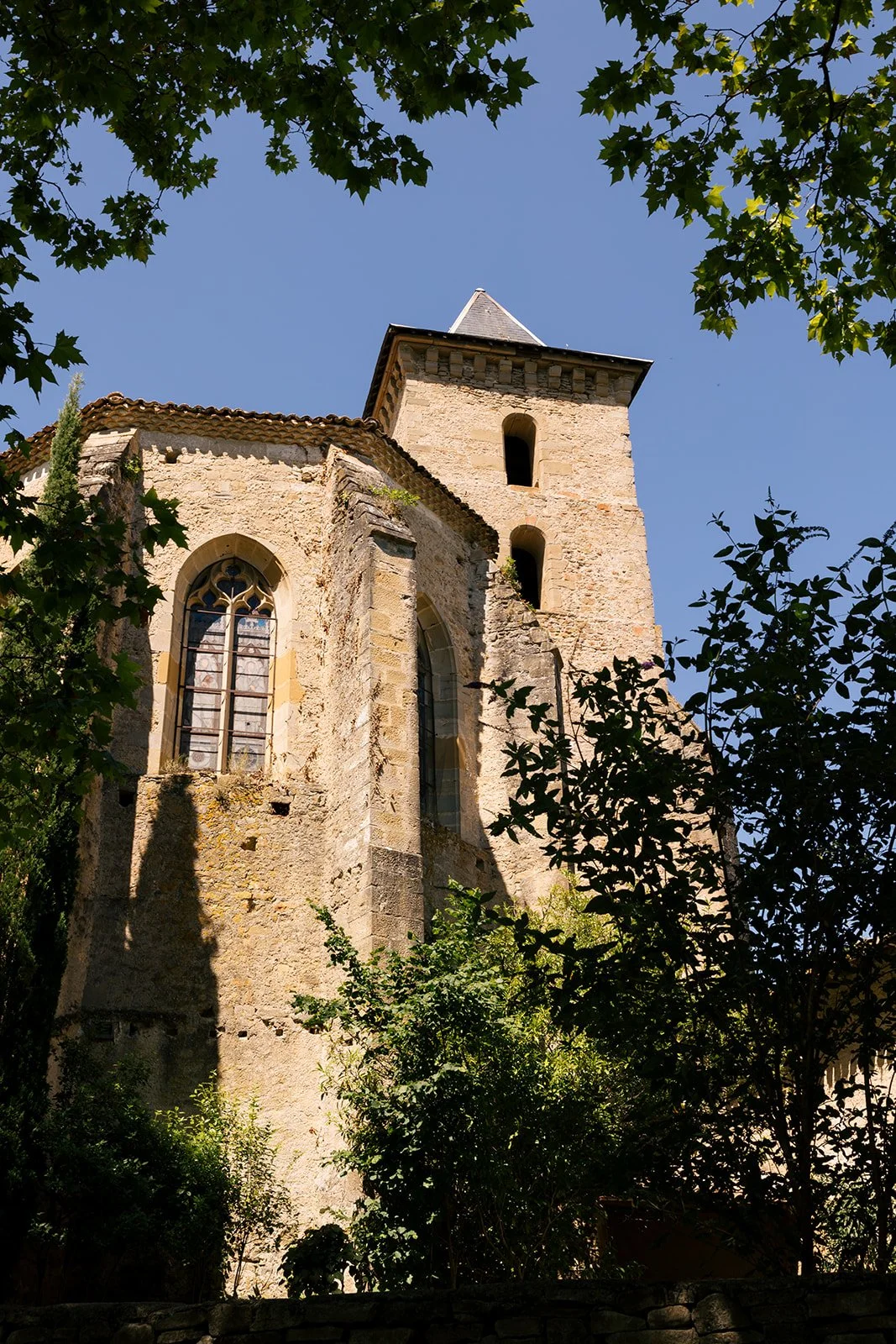 An old stone church with tall arched windows, surrounded by trees and greenery, under a clear blue sky.