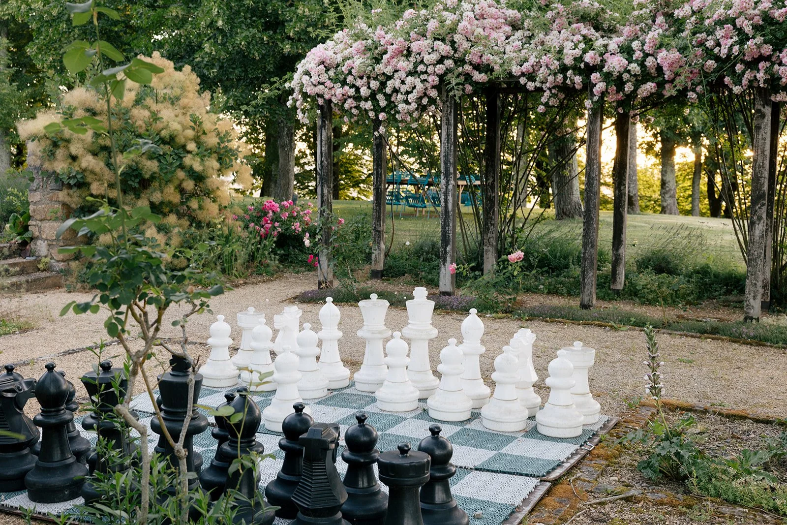 Giant outdoor chess set on a checkered tile surface in a lush garden with blooming pink and white flowers and a wooden trellis structure in the background.