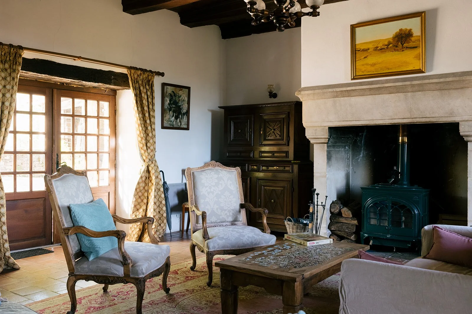 A cozy living room with vintage furniture, including two upholstered armchairs, a small wooden coffee table with a puzzle in progress, and a pink sofa partially visible. The room has a fireplace with a wood stove, wooden ceiling beams, patterned curtains by a glass door, and framed artwork on the walls.